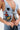 A person wears the OUT OF MY LEAGUE DENIM HALTER TOP featuring colorful floral and cartoon-style prints, paired with layered silver body jewelry and visible tattoos on their chest and hand.