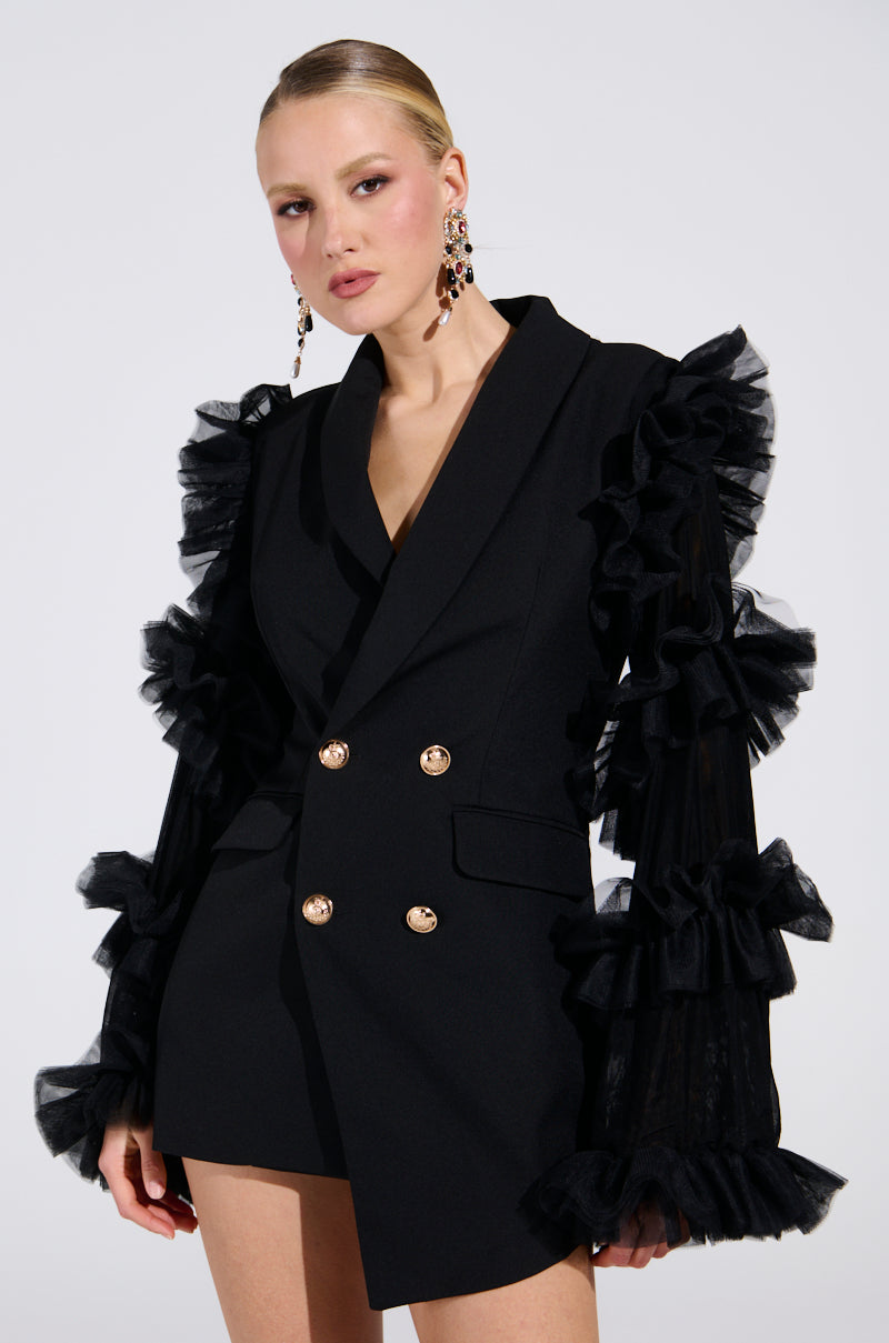 Confident in the chic LIVING OVER THE TOP RUFFLE SLEEVE BLAZER MINI DRESS, a person stands with slicked-back hair and large earrings accentuating the plunging neckline, while sheer ruffled sleeves add flair against a plain, light background that highlights their sophisticated style.