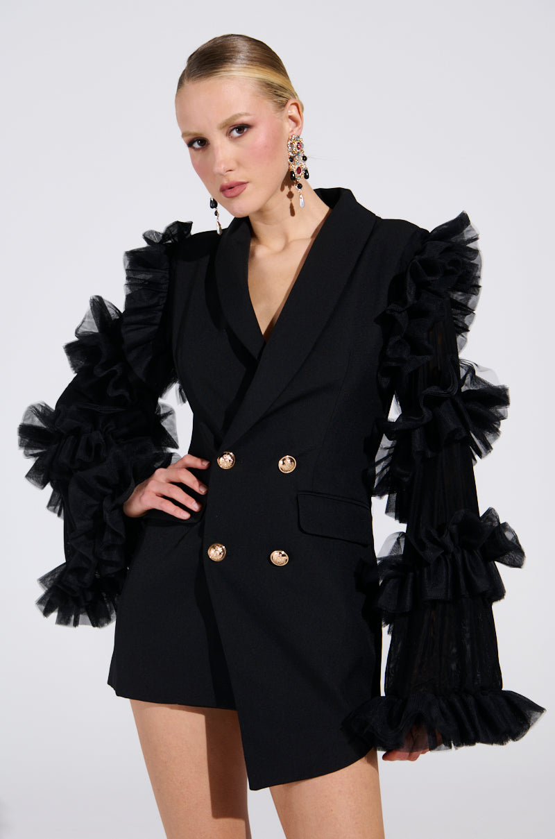 In a display of luxury fashion, someone poses confidently in the LIVING OVER THE TOP RUFFLE SLEEVE BLAZER MINI DRESS, exuding modern elegance. Their short hair and bold earrings enhance the dramatic ruffled tulle sleeves as they strike a hand-on-hip pose against a plain backdrop.