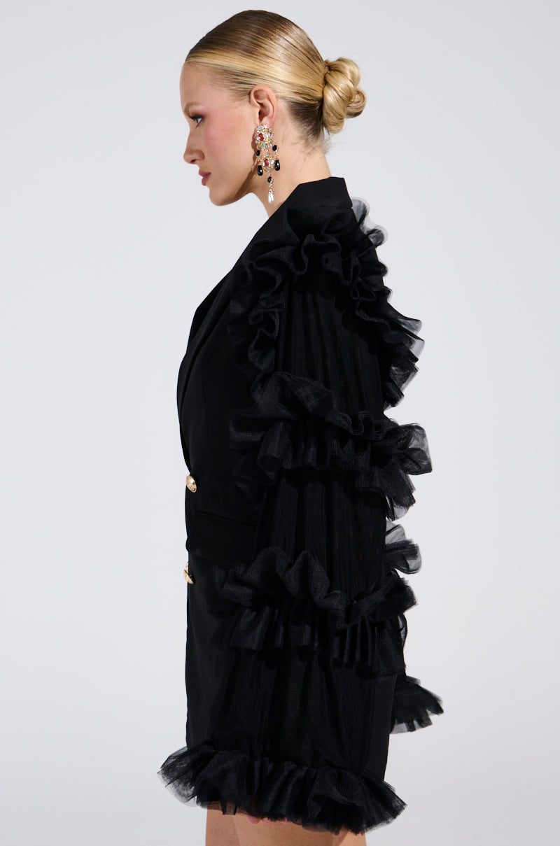 A woman in profile wears the LIVING OVER THE TOP RUFFLE SLEEVE BLAZER MINI DRESS with elegant ruffle sleeves and hem, styled with a neat bun and large, decorative earrings, exuding sophistication against a plain background.