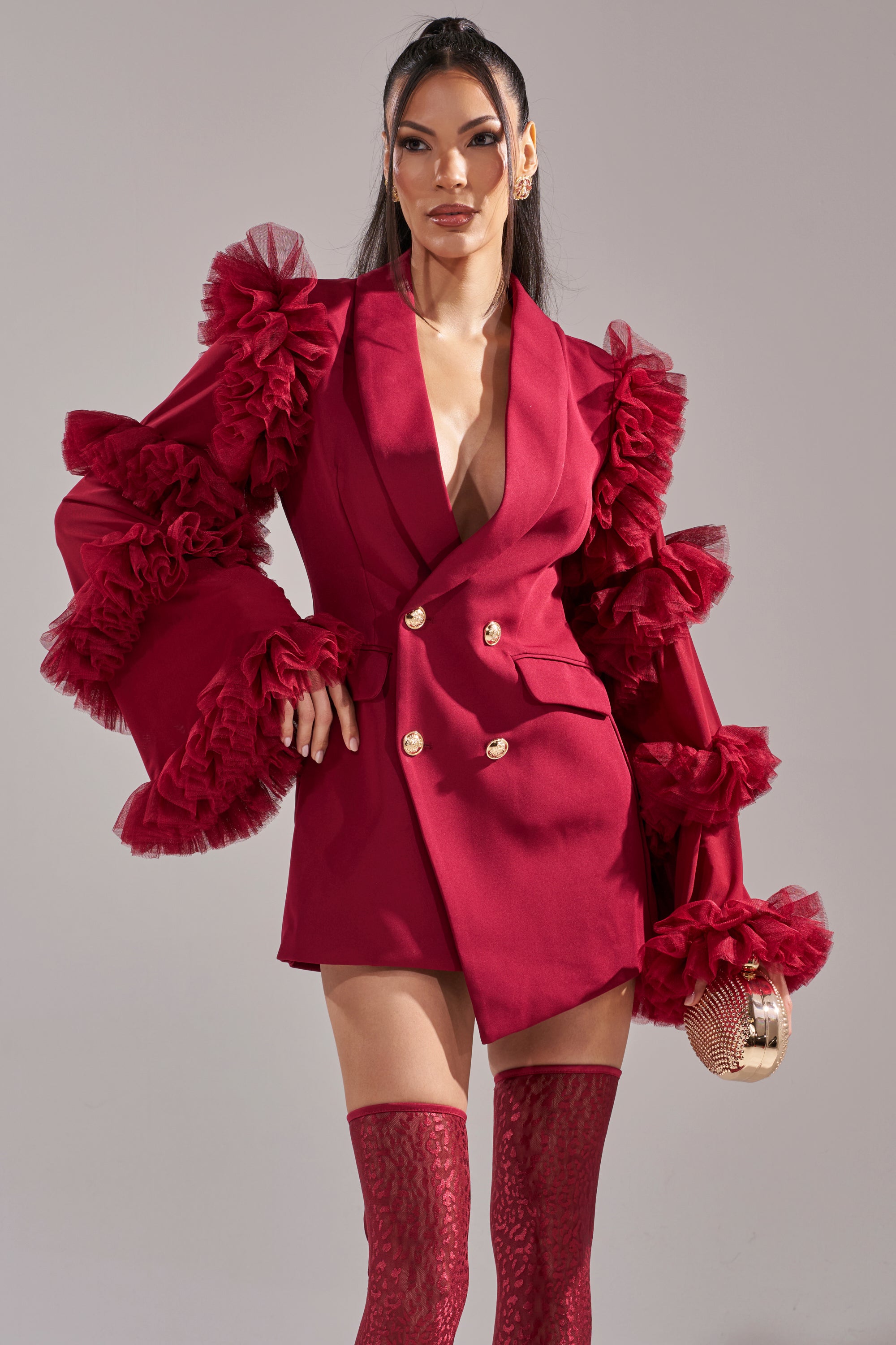 A woman exudes confidence in the LIVING OVER THE TOP RUFFLE SLEEVE BLAZER MINI DRESS IN BURGUNDY, styled with ruffled tulle sleeves, matching thigh-high stockings, gold earrings, a round clutch, and long straight hair.