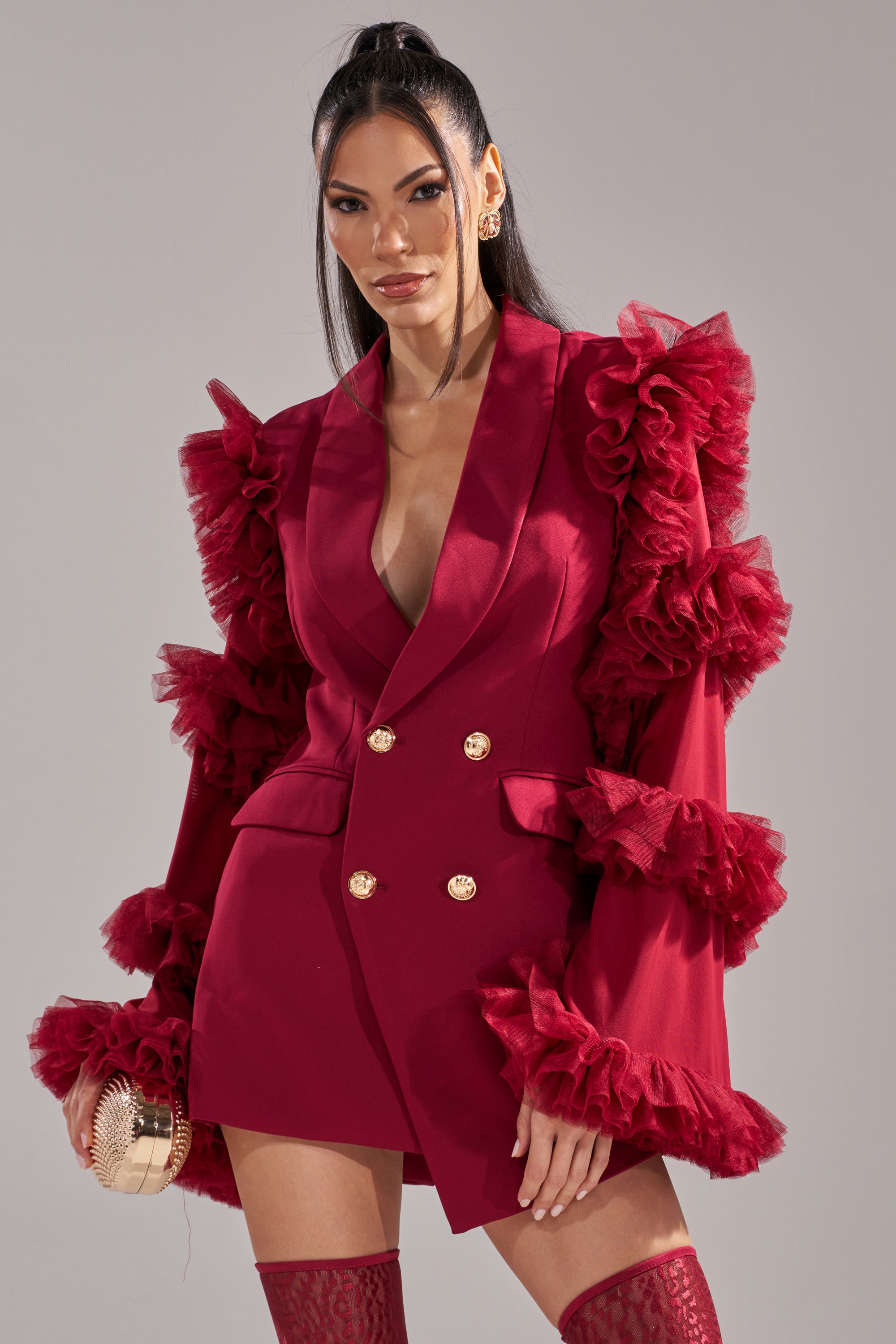 Wearing the LIVING OVER THE TOP RUFFLE SLEEVE BLAZER MINI DRESS IN BURGUNDY, a woman stands confidently with a gold clutch, red thigh-high stockings, and long dark hair styled half-up. 2
