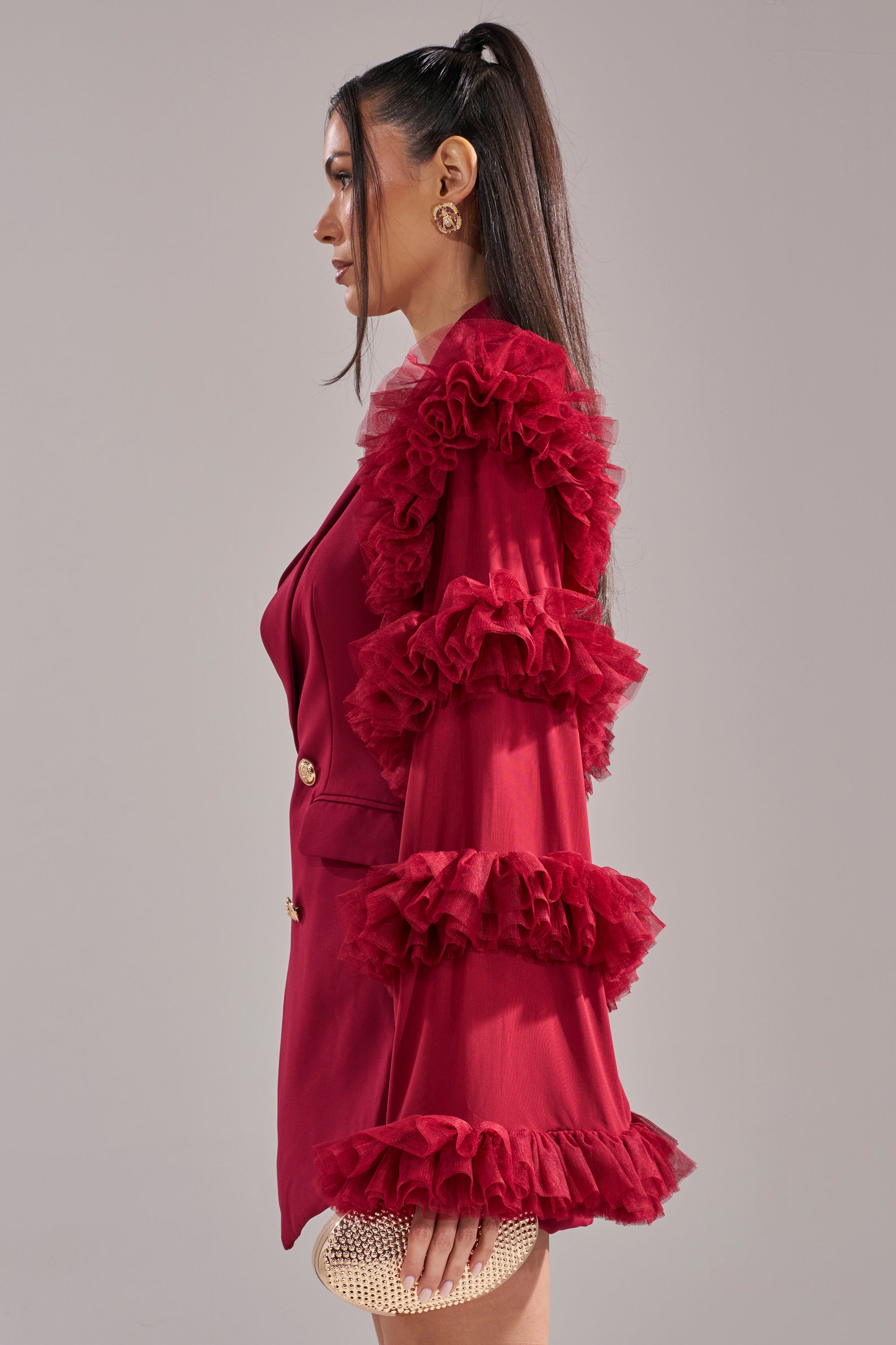 A woman with long dark hair in a ponytail wears the LIVING OVER THE TOP RUFFLE SLEEVE BLAZER MINI DRESS IN BURGUNDY. She holds a small, textured gold clutch and stands in profile against a plain background. 3