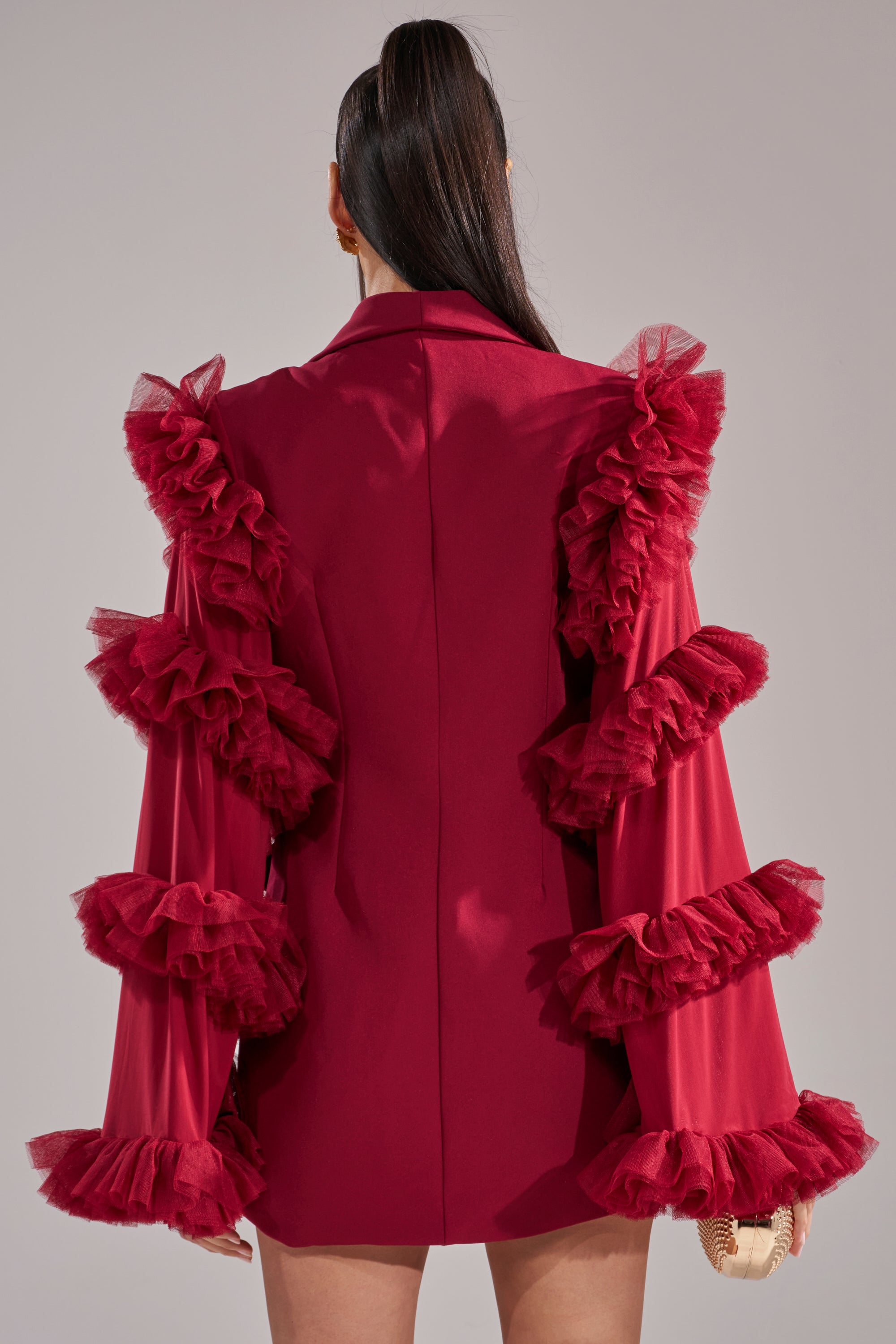 A woman with long, dark hair in a high ponytail is seen from the back wearing the LIVING OVER THE TOP RUFFLE SLEEVE BLAZER MINI DRESS IN BURGUNDY, featuring dramatic tulle sleeves and a tailored blazer silhouette, against a plain background. 4