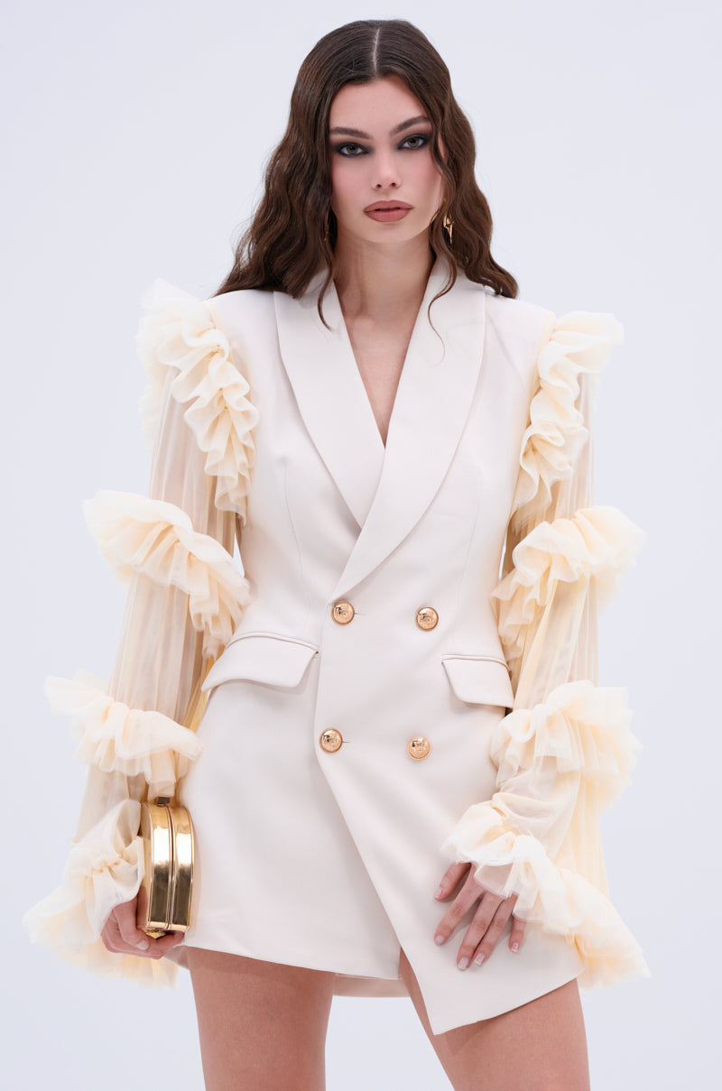 A woman with long brown hair models the LIVING OVER THE TOP RUFFLE SLEEVE BLAZER MINI DRESS, showcasing its sleek gold buttons and sheer ruffled sleeves. She holds a gold clutch and stands gracefully against a plain background.