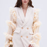 A woman with long brown hair models the LIVING OVER THE TOP RUFFLE SLEEVE BLAZER MINI DRESS, showcasing its sleek gold buttons and sheer ruffled sleeves. She holds a gold clutch and stands gracefully against a plain background.