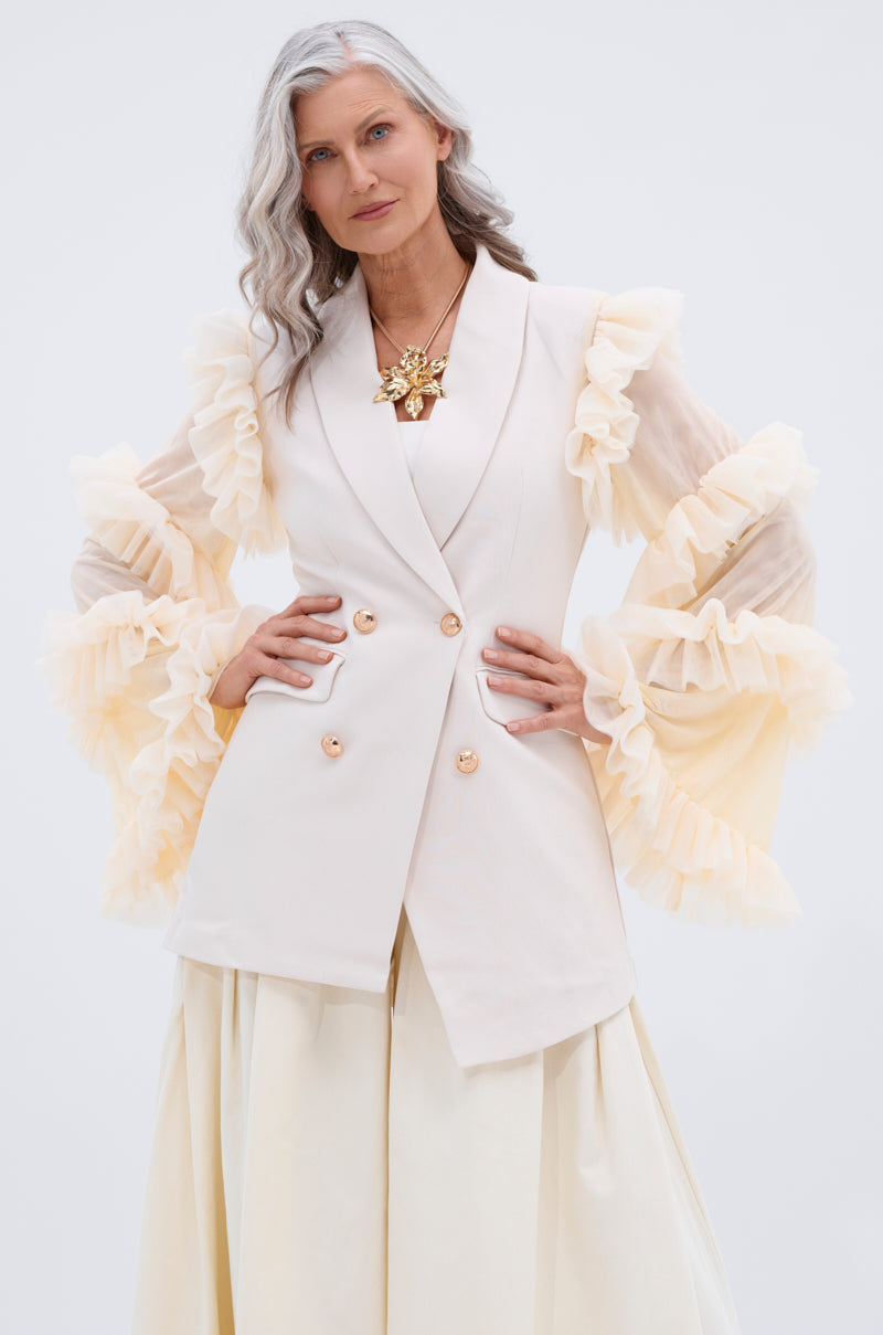 A confident older woman with long gray wavy hair models the LIVING OVER THE TOP RUFFLE SLEEVE BLAZER MINI DRESS and a bold gold pendant necklace against a plain light background.