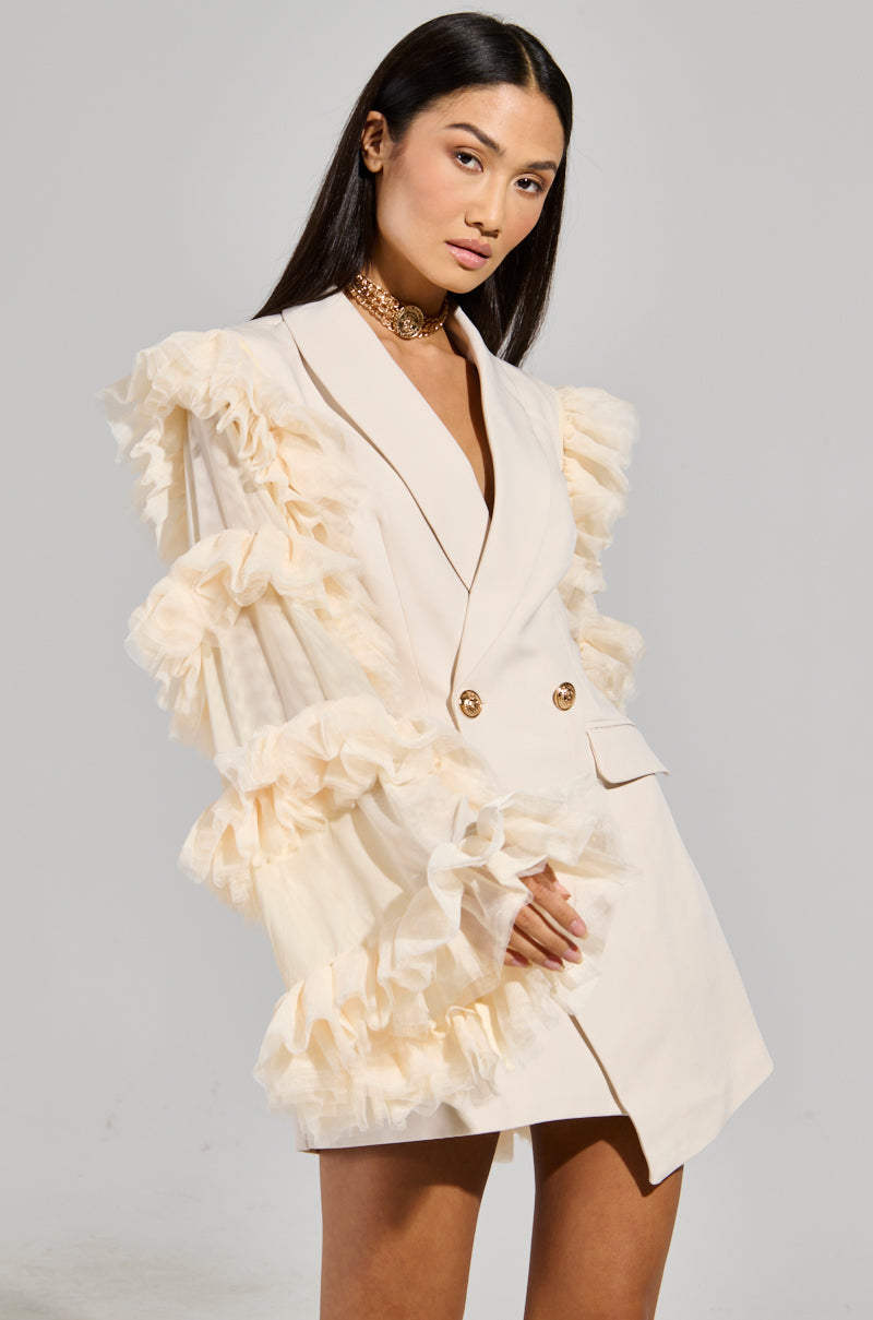 A woman poses confidently, wearing the LIVING OVER THE TOP RUFFLE SLEEVE BLAZER MINI DRESS, a chic cream-colored mini dress featuring a double-breasted front and stylish layered ruffles on the sleeves. She accessorizes with a gold choker necklace, standing against a plain white background.