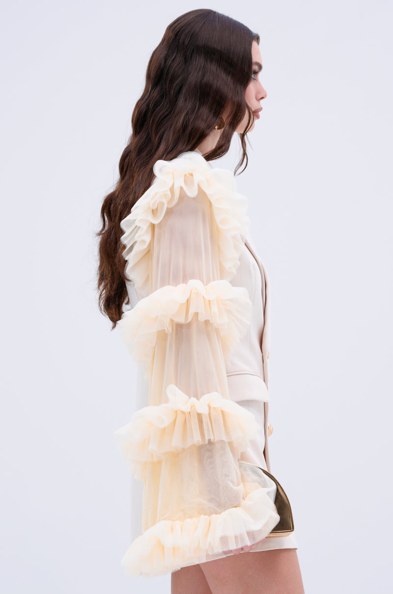 A woman with long, wavy hair is elegantly dressed in the LIVING OVER THE TOP RUFFLE SLEEVE BLAZER MINI DRESS, showcasing dramatic sheer ruffle sleeves. She poses gracefully against a plain light background.