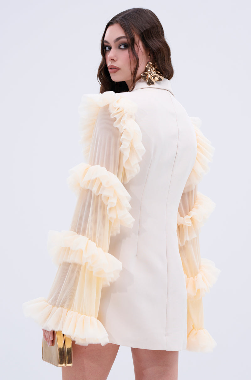 A woman with long dark hair wears the "Living Over the Top Ruffle Sleeve Blazer Mini Dress" and holds a gold clutch. She stands against a plain backdrop, looking over her shoulder.