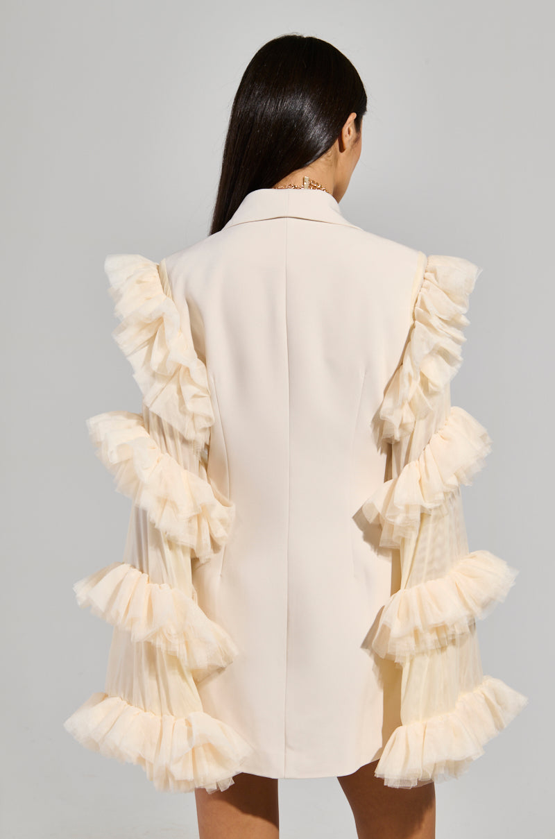 A person with long dark hair is wearing the "Living Over The Top Ruffle Sleeve Blazer Mini Dress" in pale cream, featuring layered ruffle details along the sleeves and sides. The image is taken from behind against a plain background, highlighting the garment's delicate design.