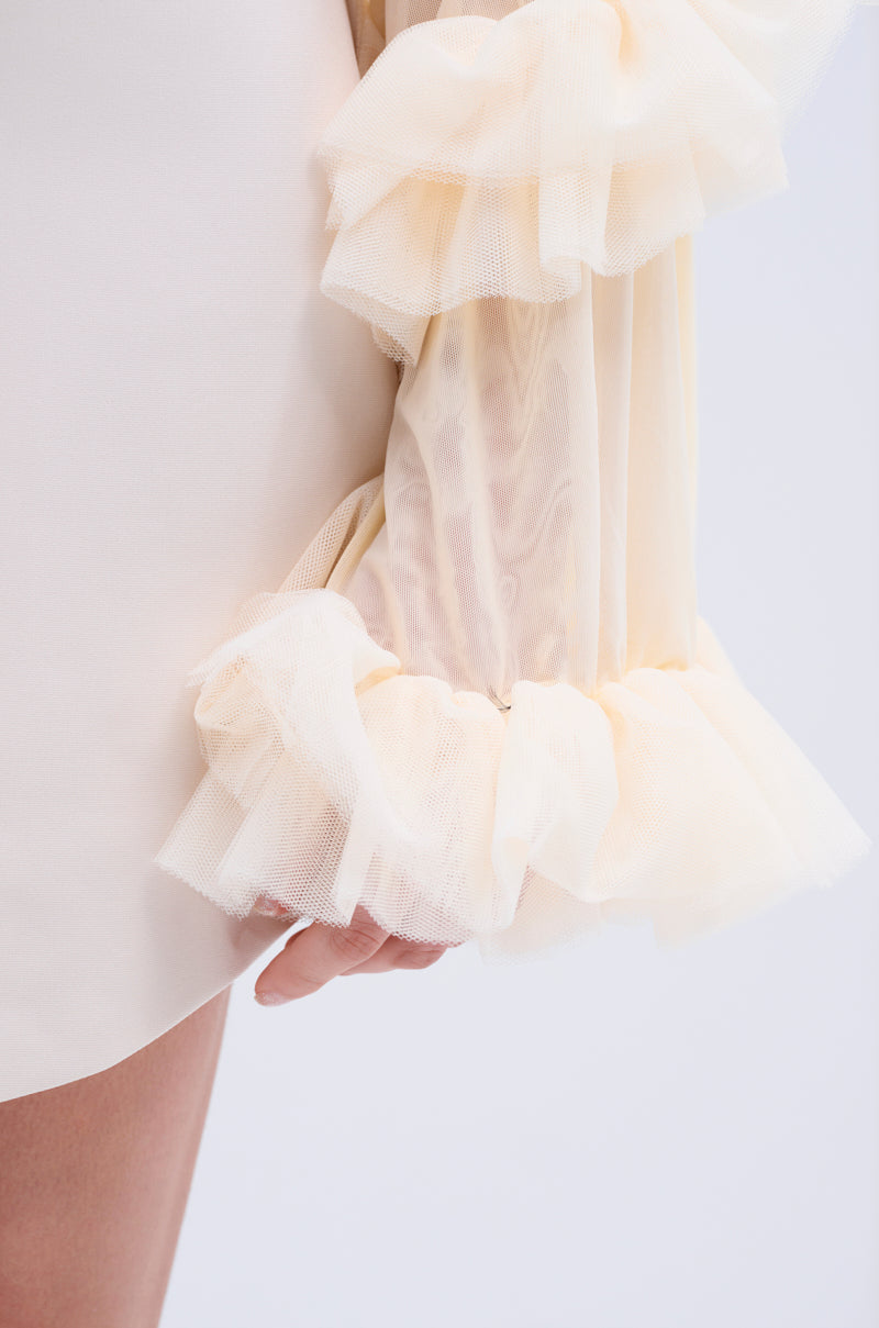 A close-up shows a person wearing the LIVING OVER THE TOP RUFFLE SLEEVE BLAZER MINI DRESS, featuring sheer, ruffled tulle layers on the sleeves that reveal a hand beneath. The light, delicate cream fabric stands out against a plain, light-colored background, enhancing its elegance.