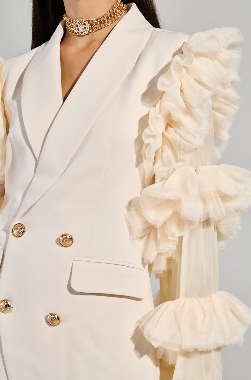 A person wears the chic LIVING OVER THE TOP RUFFLE SLEEVE BLAZER MINI DRESS, featuring a stylish cream-colored design with dramatic tulle accents and gold buttons. This double-breasted piece with a sharp collar is perfectly paired with a gold statement choker necklace.