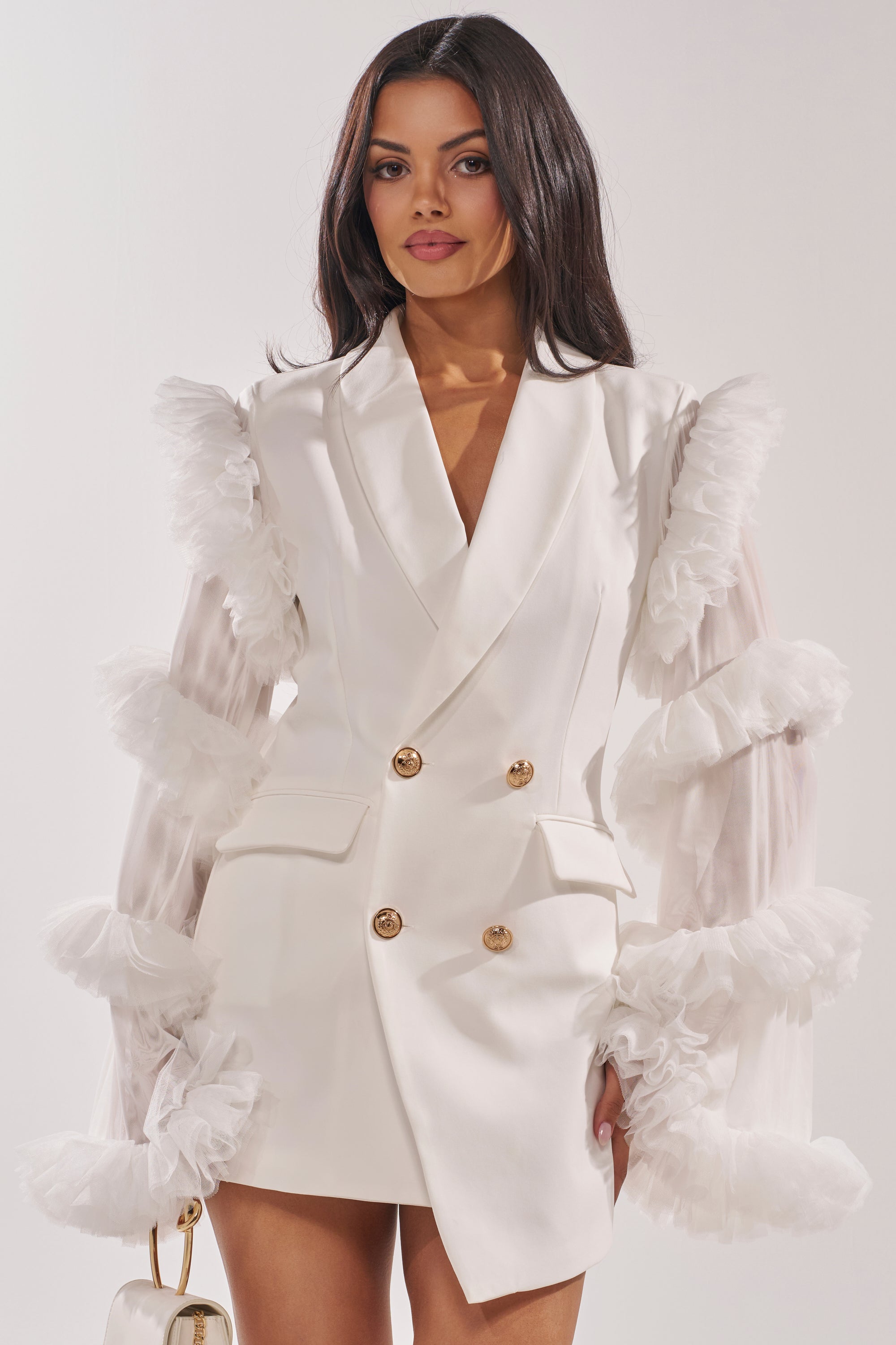 A woman poses confidently against a plain light background in the LIVING OVER THE TOP RUFFLE SLEEVE BLAZER MINI DRESS IN WHITE, featuring gold buttons and tulle ruffle sleeves. She holds a small white handbag to complete the chic look. 0