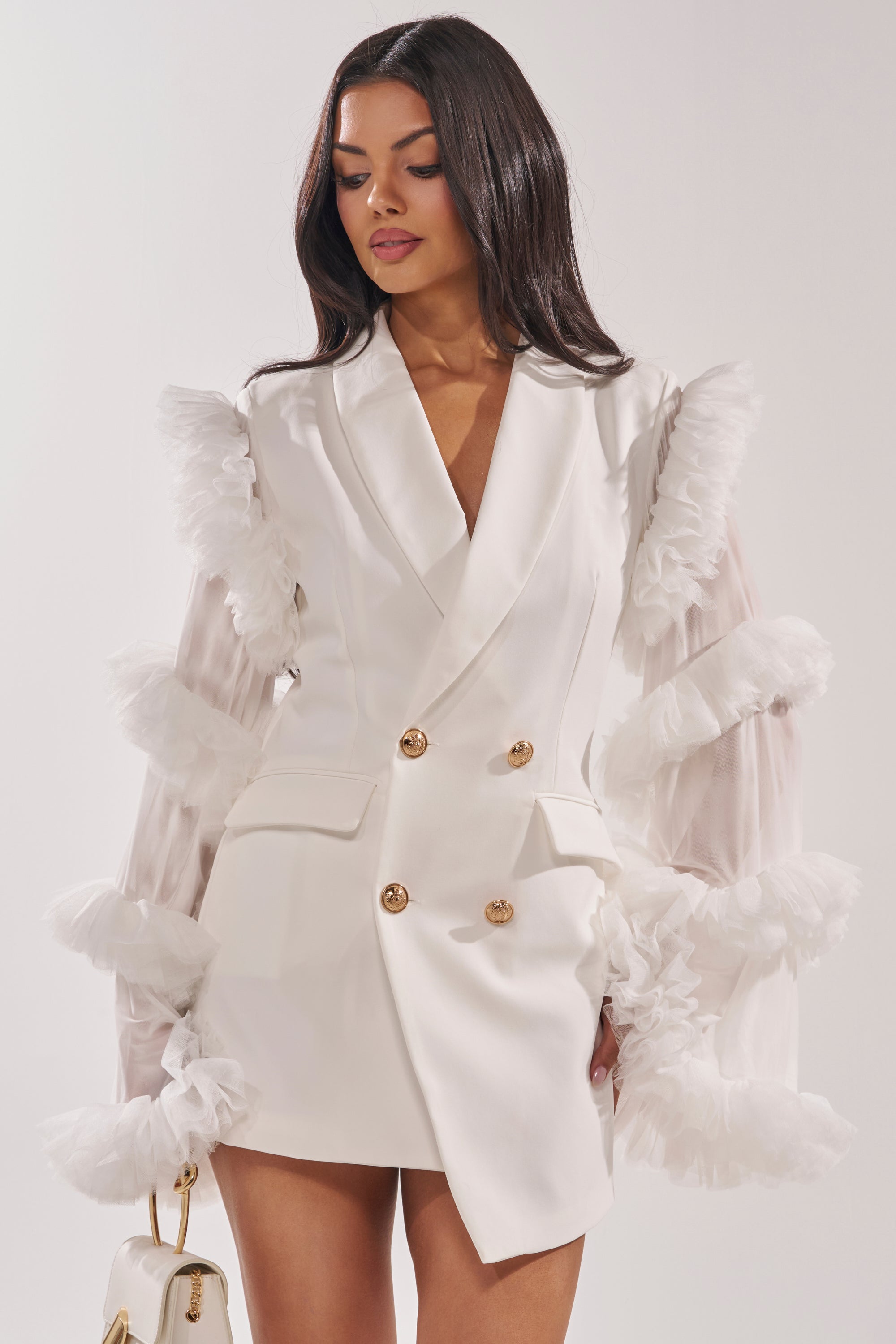 A woman wears the LIVING OVER THE TOP RUFFLE SLEEVE BLAZER MINI DRESS IN WHITE, a tailored mini dress with gold buttons and sheer ruffled tulle sleeves. She carries a small white handbag, her long dark hair styled in soft waves. 1