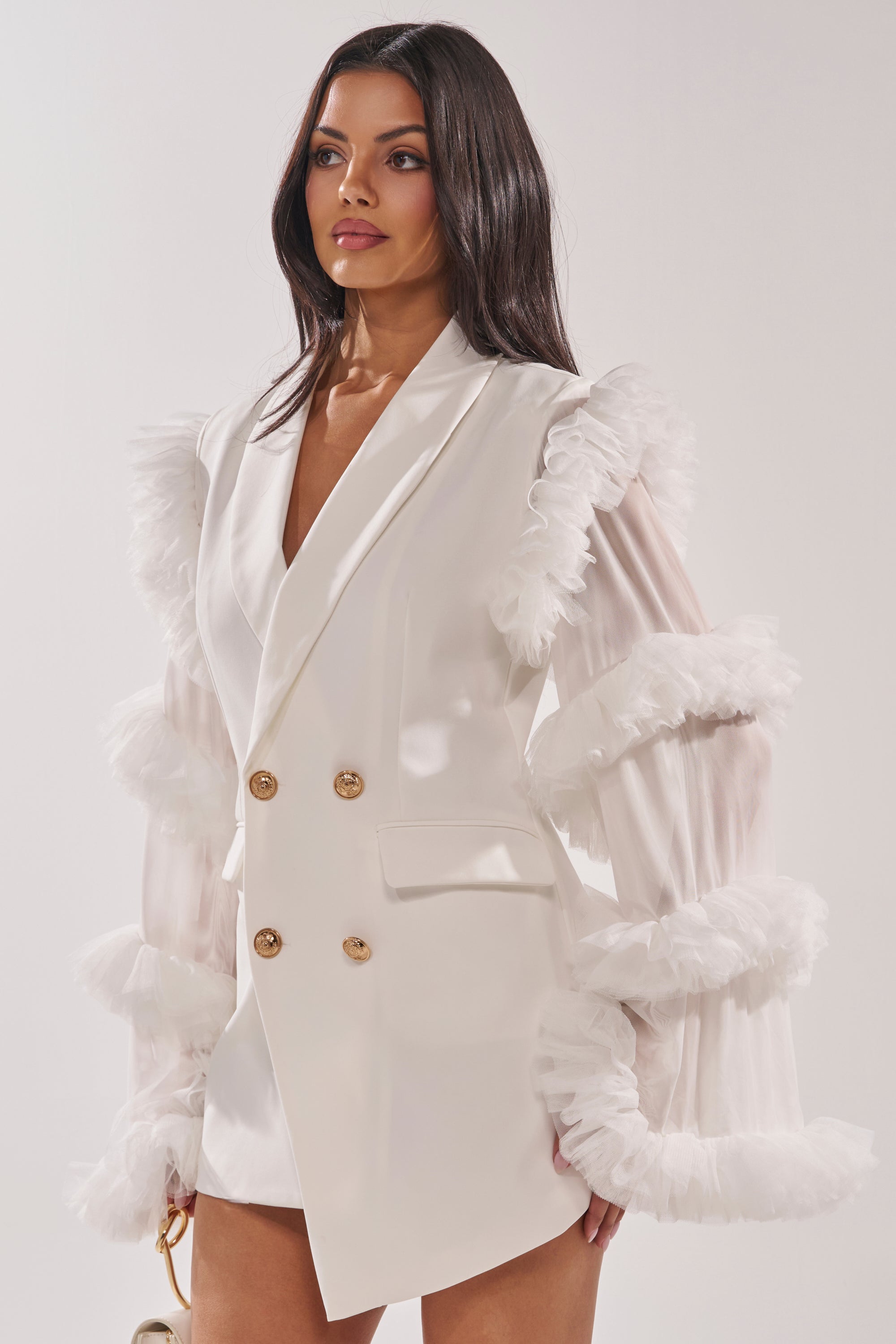 A woman stands against a plain white background wearing the LIVING OVER THE TOP RUFFLE SLEEVE BLAZER MINI DRESS IN WHITE, featuring tulle ruffle sleeves and gold buttons, and holds a small gold handbag. 2