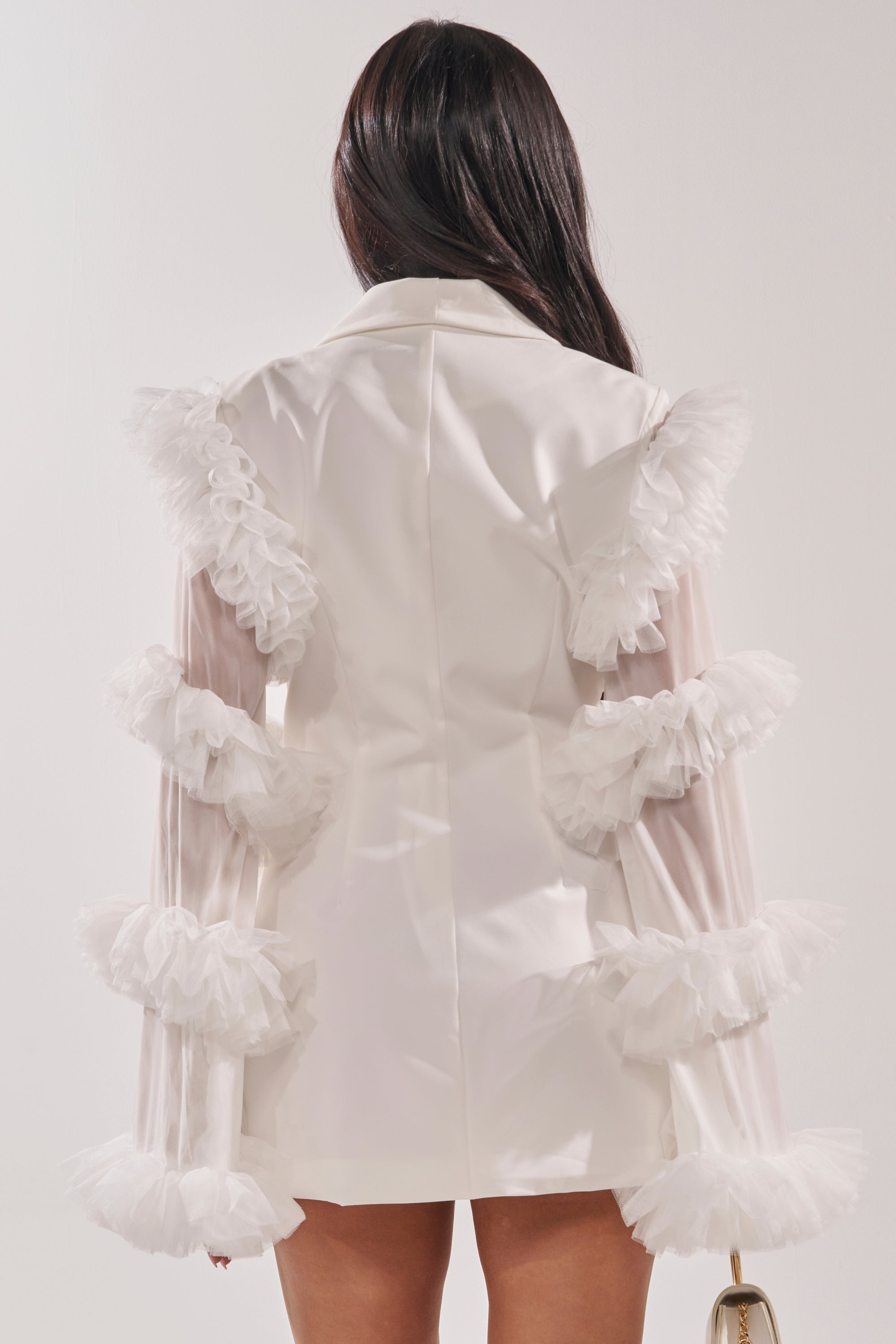 A woman faces away, wearing the LIVING OVER THE TOP RUFFLE SLEEVE BLAZER MINI DRESS IN WHITE with sheer tulle ruffle sleeves and a tailored silhouette, holding a small gold handbag against a plain white background. 3