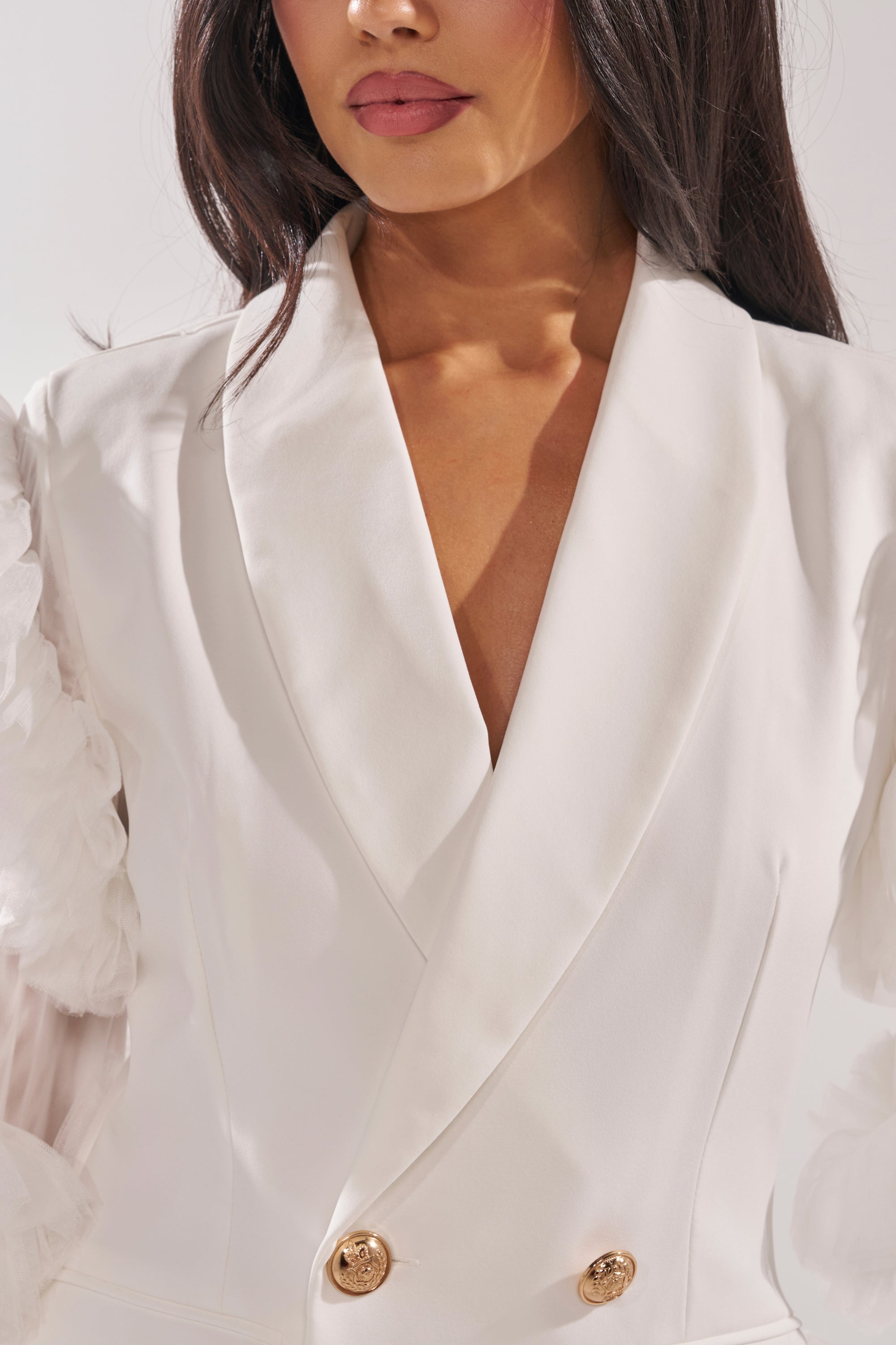 Wearing the LIVING OVER THE TOP RUFFLE SLEEVE BLAZER MINI DRESS IN WHITE, featuring tulle ruffle sleeves, gold buttons, and a double-breasted design, a woman with loose dark waves is shown from shoulders to mid-torso. 6