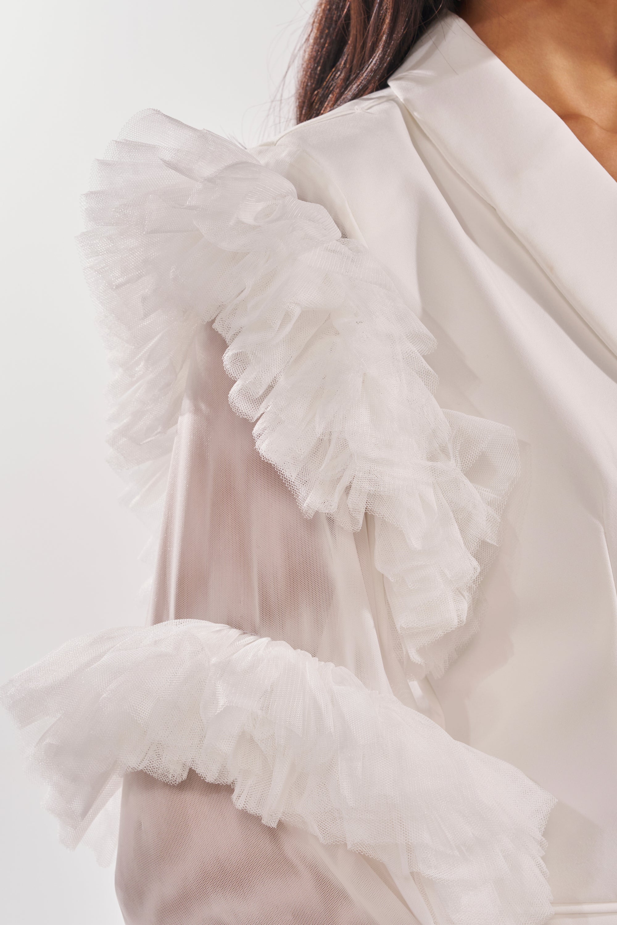 Close-up of the LIVING OVER THE TOP RUFFLE SLEEVE BLAZER MINI DRESS IN WHITE, featuring layered tulle ruffle sleeves on the shoulders and upper arm, worn by a person with long dark hair against a plain light background. 8