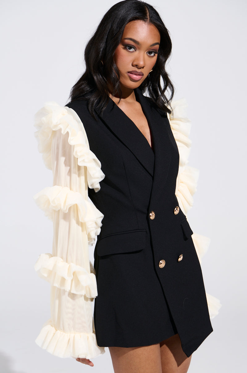 Against a plain backdrop, a woman wearing the LIVING OVER THE TOP RUFFLE SLEEVE BLAZER MINI DRESS in black, featuring gold buttons and cream ruffled sleeves, gazes slightly to the side with her hair styled in soft waves.