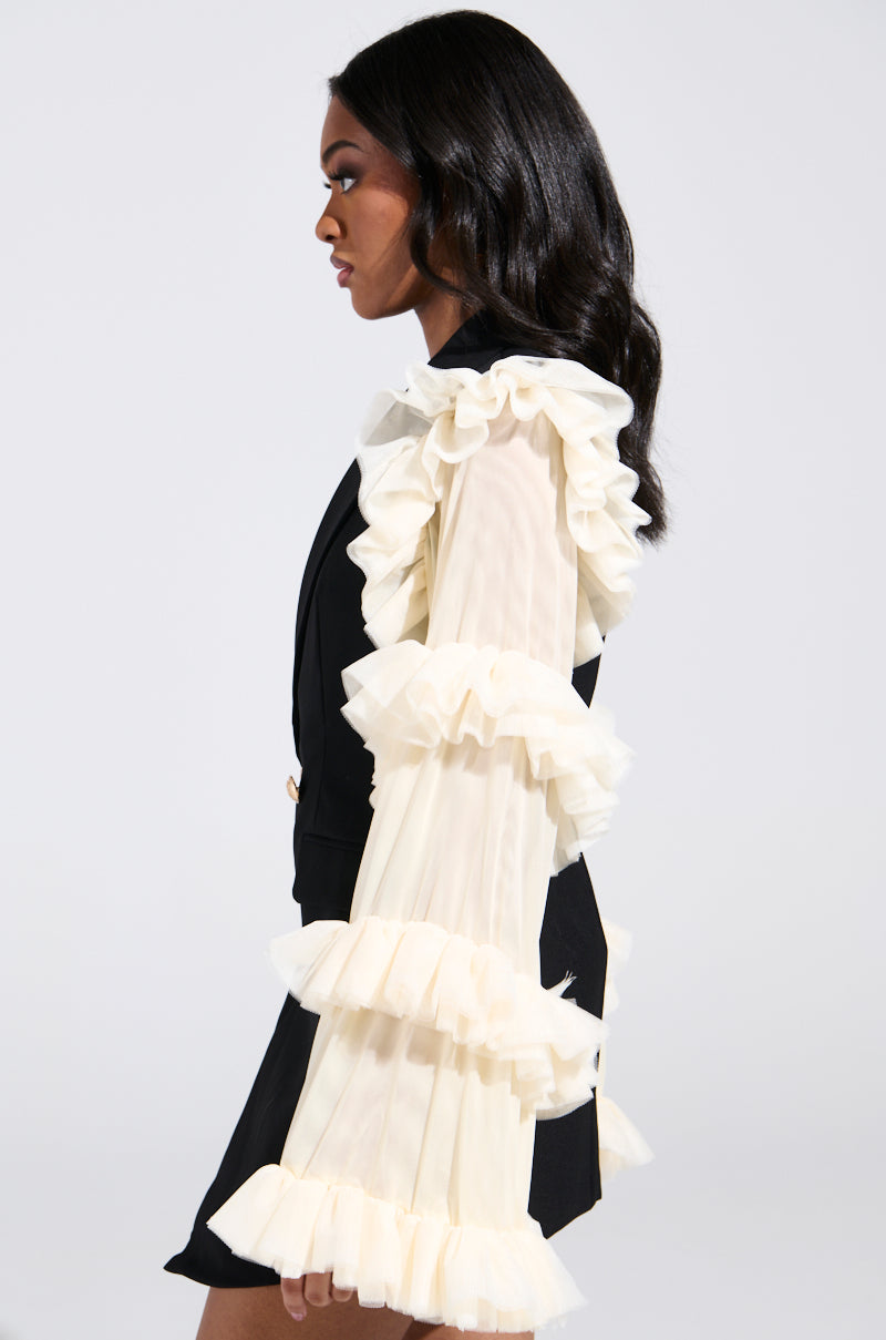 A woman in profile dons a chic outfit featuring a black vest with the LIVING OVER THE TOP RUFFLE SLEEVE BLAZER MINI DRESS in beige. Her dark, wavy hair complements the plain background, highlighting her stylish ensemble.
