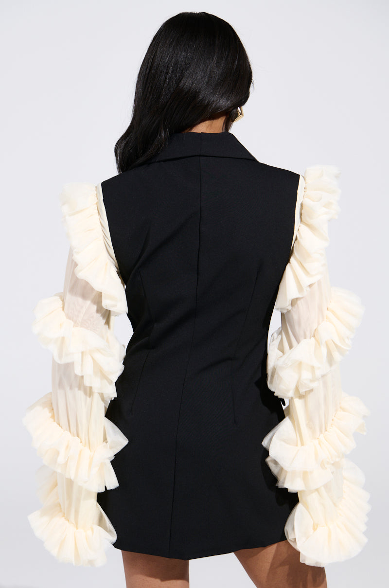Against a light gray backdrop, someone with long dark hair wears the black "LIVING OVER THE TOP RUFFLE SLEEVE BLAZER MINI DRESS," showcasing dramatic sheer beige ruffles cascading down the arms.