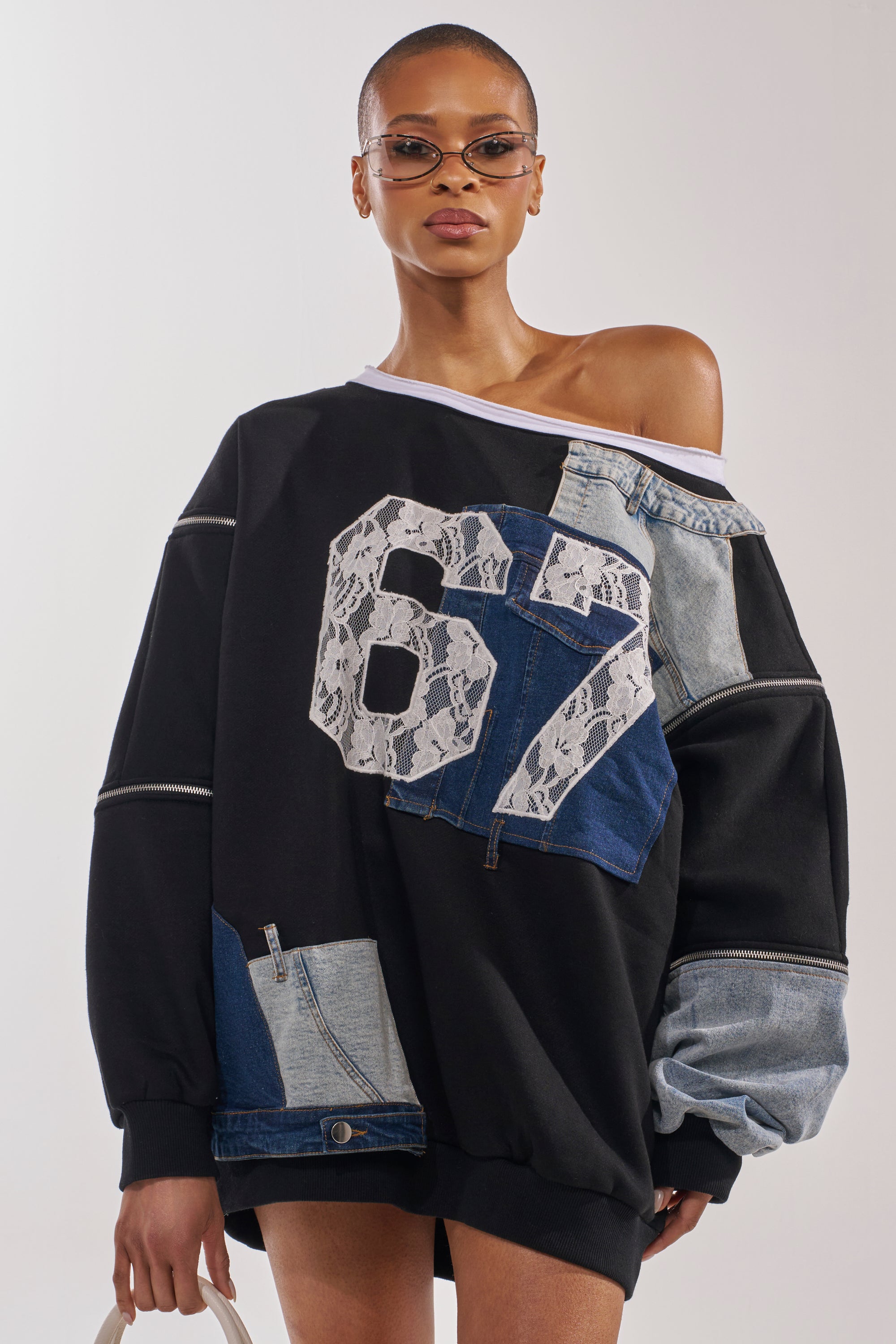 A woman wears the DREAM TEAM FRENCH TERRY MINI DRESS, an oversized off-the-shoulder sweatshirt with denim patches and a bold "67" lace detail on the front, paired with small sunglasses as she stands confidently against a plain background. 1