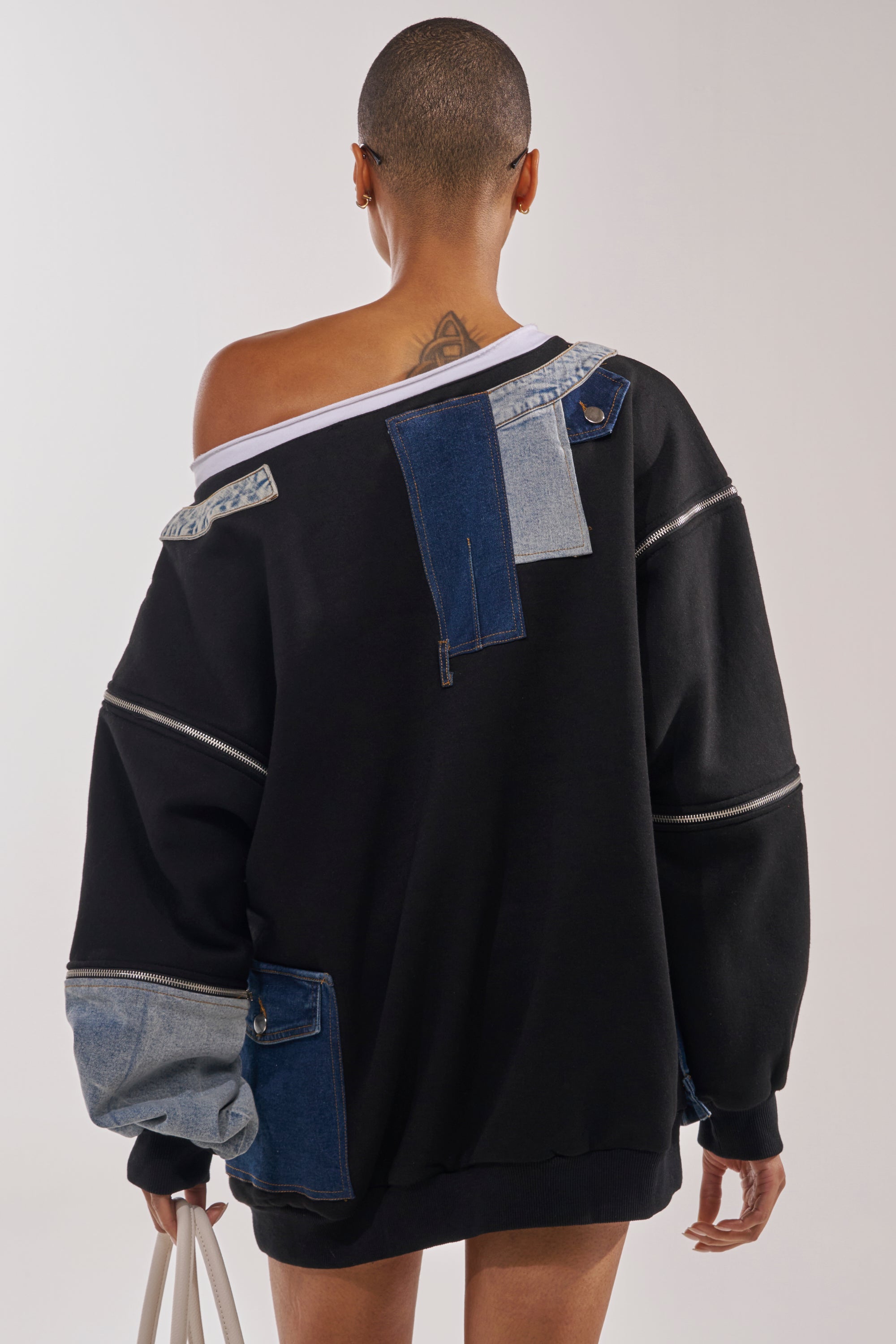 A person with a shaved head, seen from behind, wears the DREAM TEAM FRENCH TERRY MINI DRESS—an oversized off-the-shoulder style with denim patchwork. They carry a white bag and reveal an upper back tattoo. 4