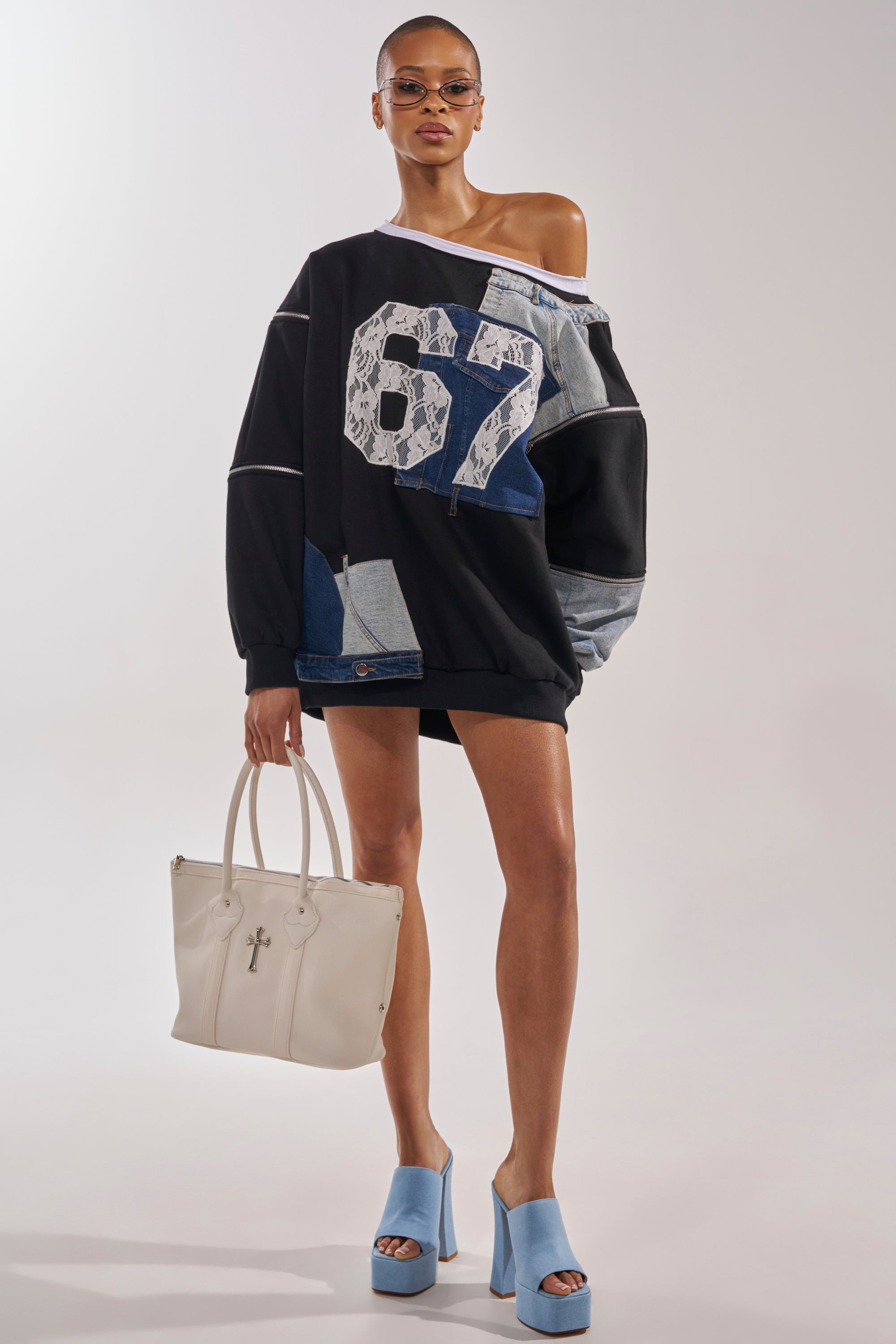 A woman in sunglasses stands against a plain background, wearing the DREAM TEAM FRENCH TERRY MINI DRESS—an oversized black sweatshirt dress with "67" and denim patches—paired with short black shorts, light blue platform heels, and a white handbag. 5