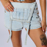 A person wearing the SELFISH SOUL DENIM SHORT in light wash denim, featuring a unique front flap design and metal button details. The high-rise fit accentuates the tattoo of a skull on their upper thigh. They complete the look with a matching denim top and black boots against a plain gray background.