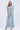A woman with long, wavy gray hair stands confidently in the COOL GIRL WIDE LEG DENIM JUMPSUIT with button front and ruffled shoulders, styled with heeled sandals against a plain white background—a true cool girl vibe.