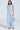 Against a plain backdrop, someone models the COOL GIRL WIDE LEG DENIM JUMPSUIT, showcasing its paneled construction with short sleeves and wide-leg pants. They accessorize with a transparent handbag and elegant silver high-heeled shoes.
