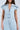 A person in the COOL GIRL WIDE LEG DENIM JUMPSUIT, a light wash cotton design with a button-up front and short sleeves. It has a fitted waist, decorative shoulder pleats, set against a simple gray background.
