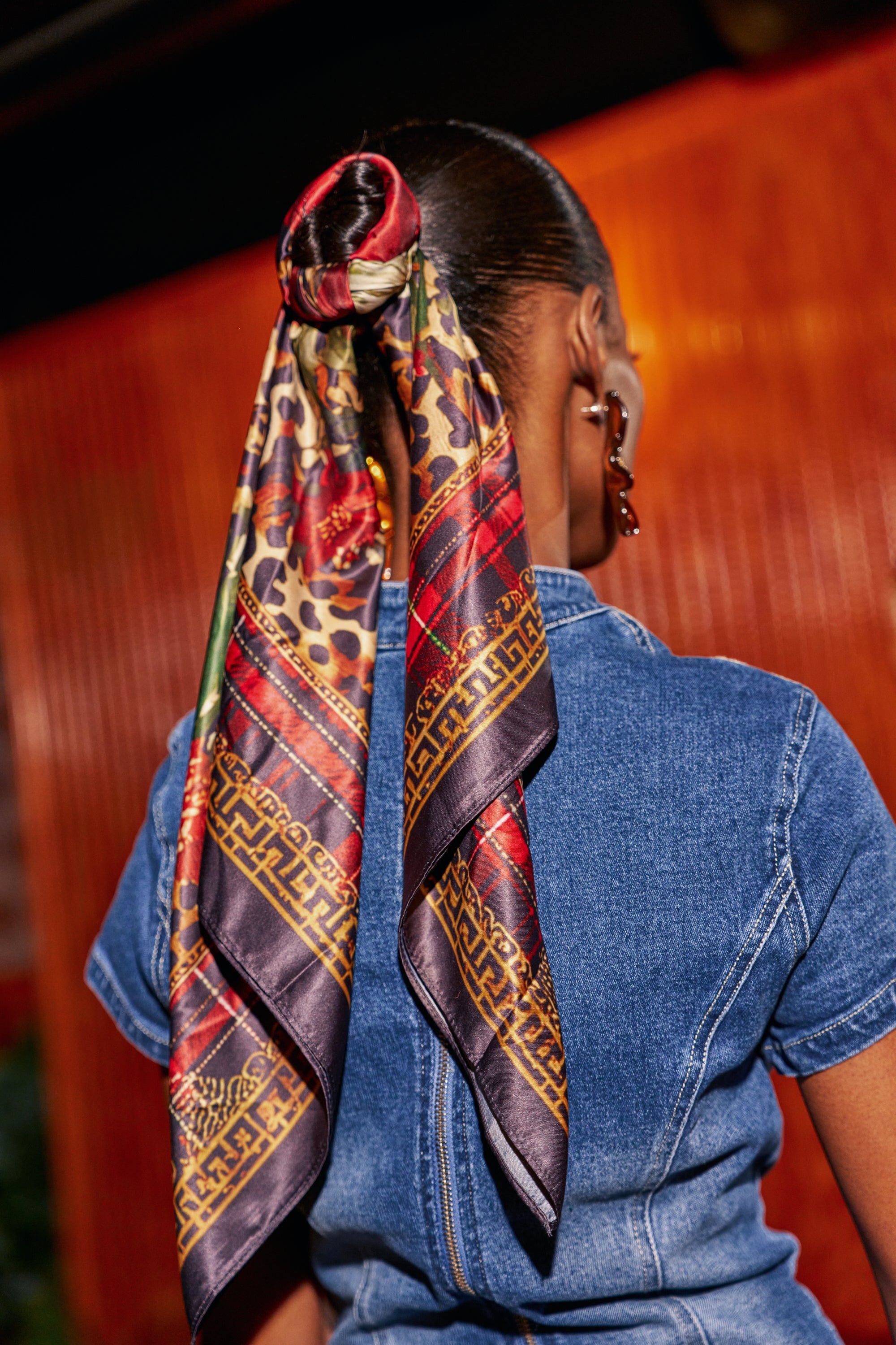 A person seen from behind wears a MIXED UP SATIN SCARF tied around a ponytail, featuring a mix of leopard print, red plaid, and floral motifs within a gold Greek-key border.