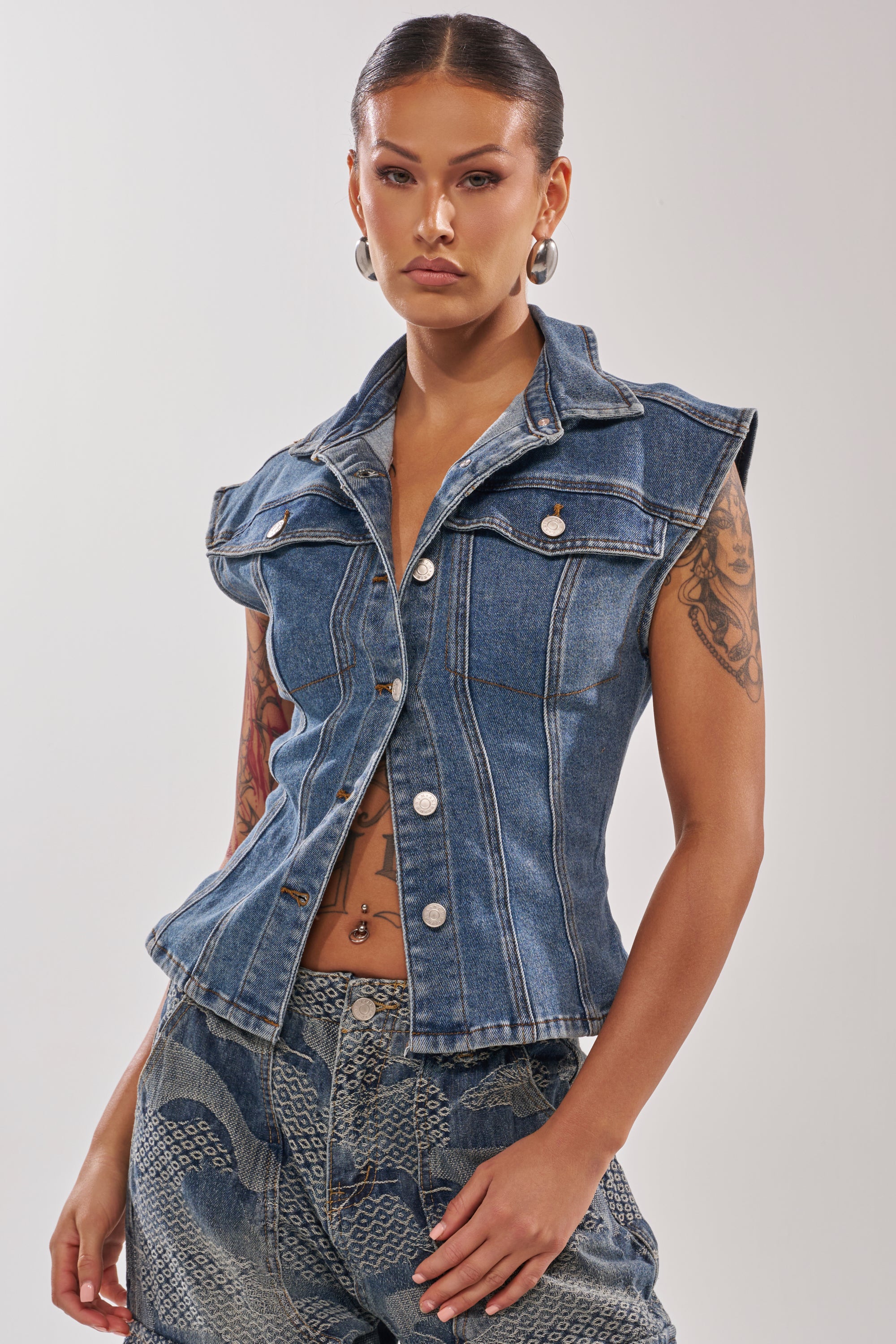 A woman with slicked-back hair wears the LILY DENIM BUTTON DOWN IN MEDIUM BLUE DENIM, styled sleeveless with silver buttons and a lace-up back, paired with matching blue patterned jeans. She accessorizes with hoop earrings and displays arm tattoos.