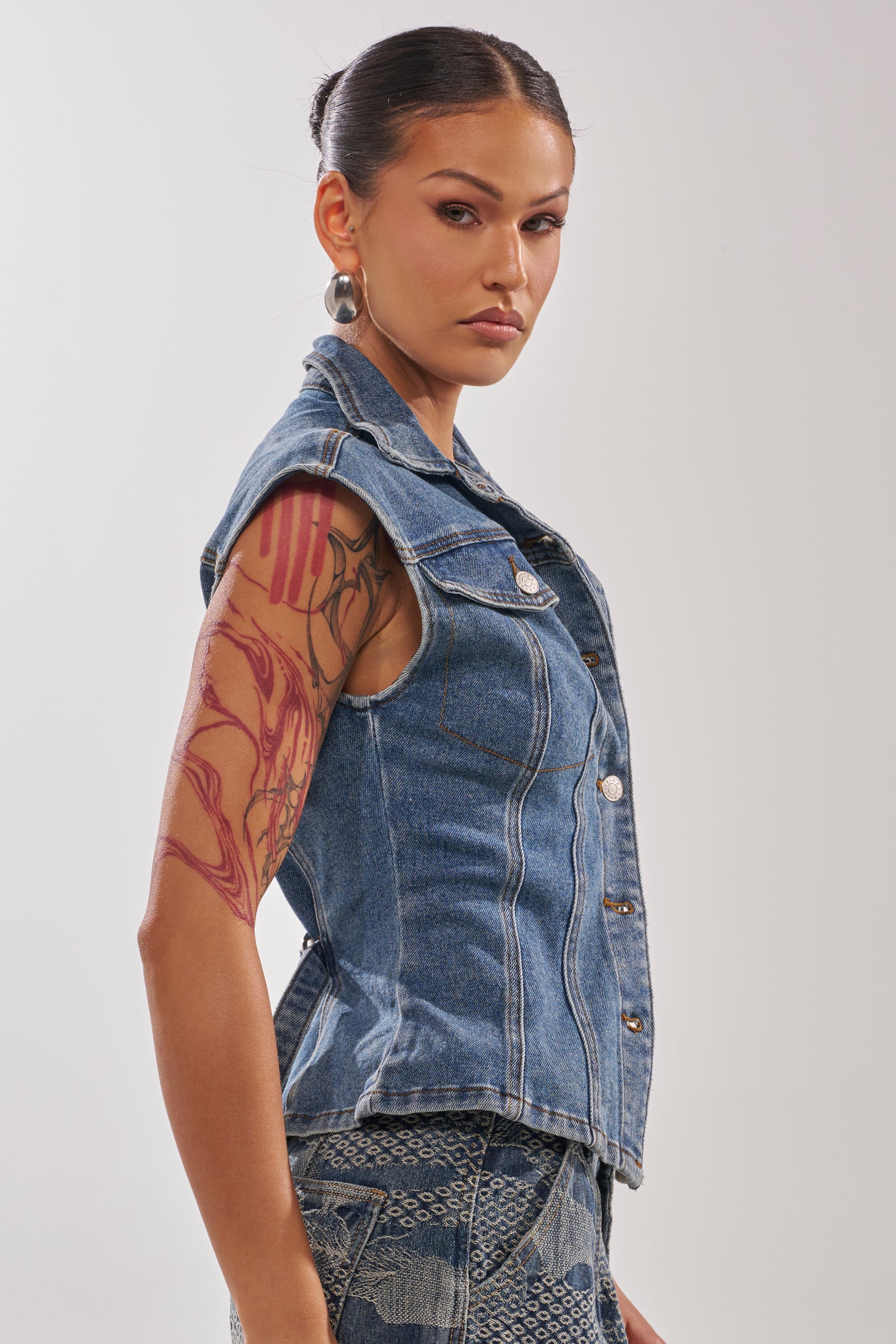 A woman with a tattooed arm wears the LILY DENIM BUTTON DOWN IN MEDIUM BLUE DENIM and patterned jeans, standing sideways against a plain light background and looking at the camera with a neutral expression.