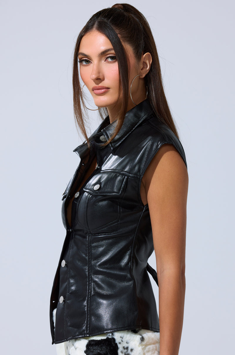 A woman with long brown hair in a high ponytail models the GUESS WHO FAUX LEATHER BUTTON DOWN, a sleeveless vest with silver snaps and front pockets, paired with black and white patterned pants against a plain light background.