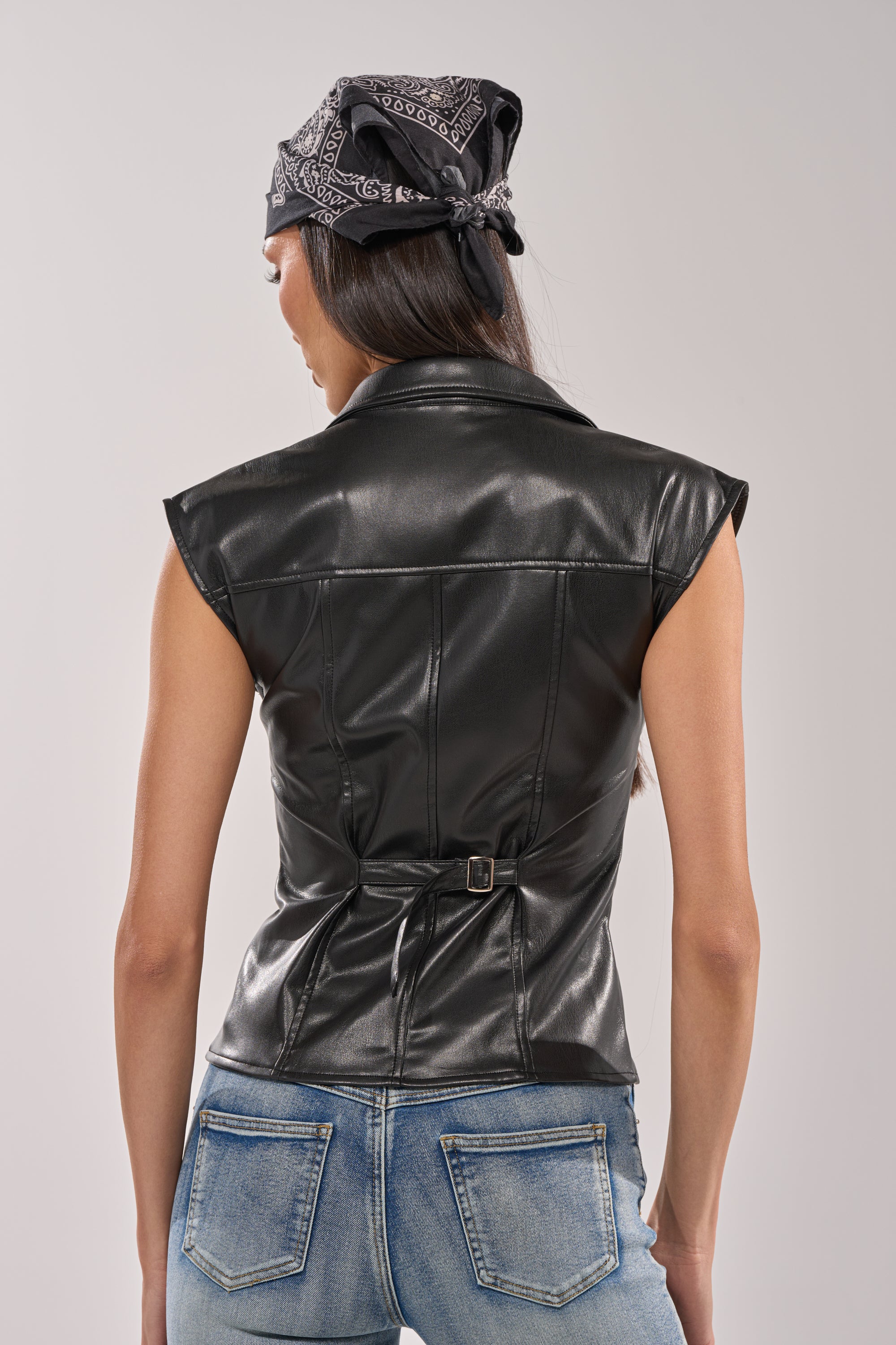 A woman seen from behind wears the GUESS WHO FAUX LEATHER BUTTON DOWN top, light blue jeans, and a black bandana tied around her head, standing against a plain light gray background. 4