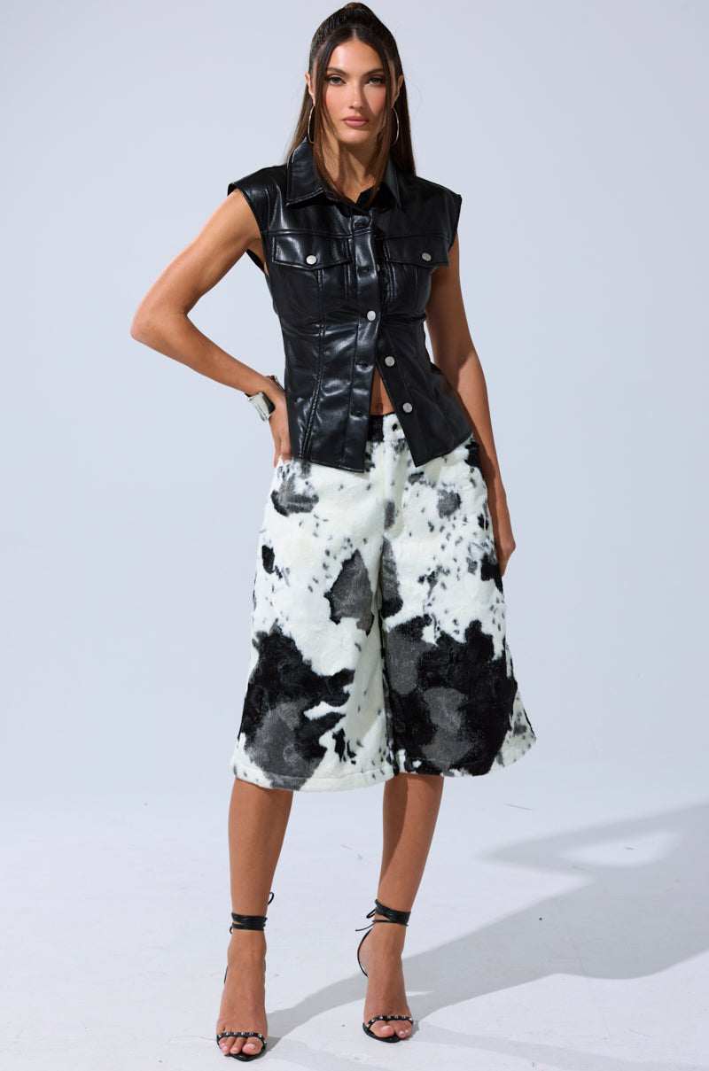 A woman stands confidently in the GUESS WHO FAUX LEATHER BUTTON DOWN, paired with wide, knee-length black-and-white abstract shorts and strappy black heels against a plain background.