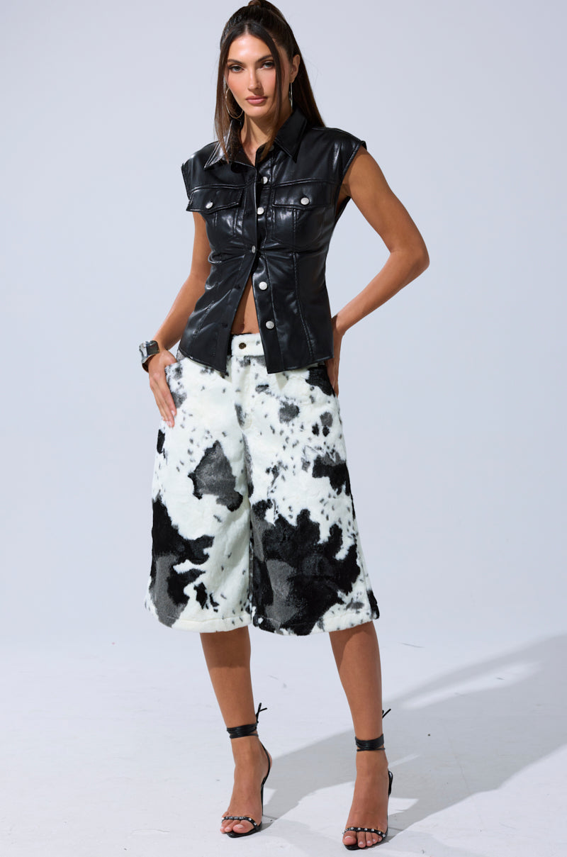 A woman wears the GUESS WHO FAUX LEATHER BUTTON DOWN, styled with knee-length cow print shorts, black high-heeled sandals, and long, straight hair.