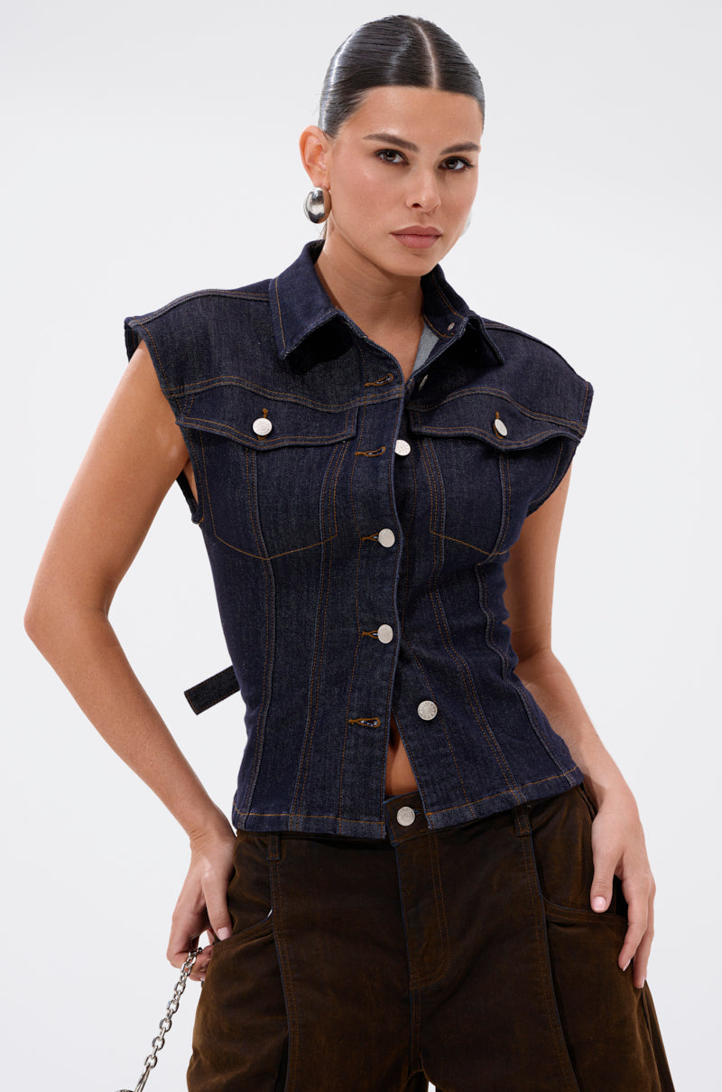 A woman with slicked-back hair models the LILY DENIM BUTTON DOWN, a sleeveless dark-wash denim top, paired with dark brown pants. She stands confidently, one hand on her hip, against a plain white background. 3