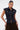 A woman with slicked-back hair models the LILY DENIM BUTTON DOWN, a sleeveless dark-wash denim top, paired with dark brown pants. She stands confidently, one hand on her hip, against a plain white background.