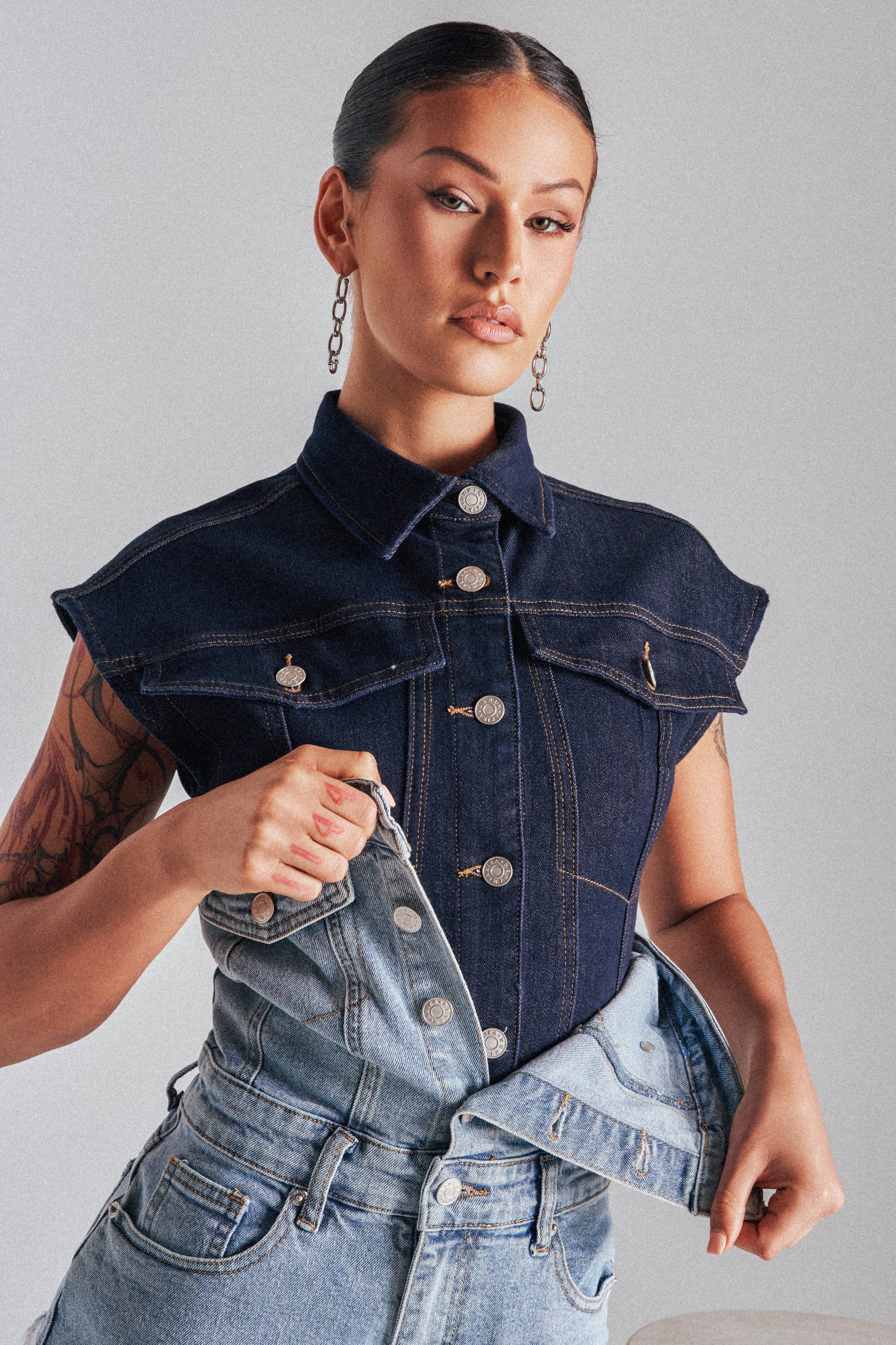A woman with slicked-back hair and hoop earrings confidently poses against a plain background, wearing the LILY DENIM BUTTON DOWN and holding a pair of light blue jeans in front of her. 0