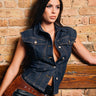 A woman with long dark hair poses against a brick wall, wearing the LILY DENIM BUTTON DOWN and black leather shorts with a brown belt. She gazes to the side with a serious expression.