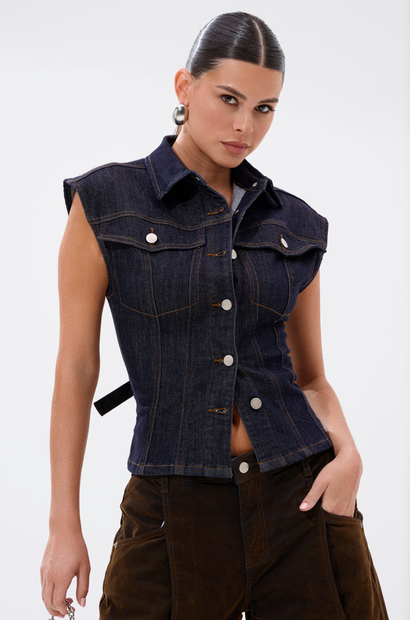 A woman with slicked-back dark hair wears the LILY DENIM BUTTON DOWN, a sleeveless dark-wash denim top, paired with brown pants. She poses against a plain white background with one hand in her pocket. 4