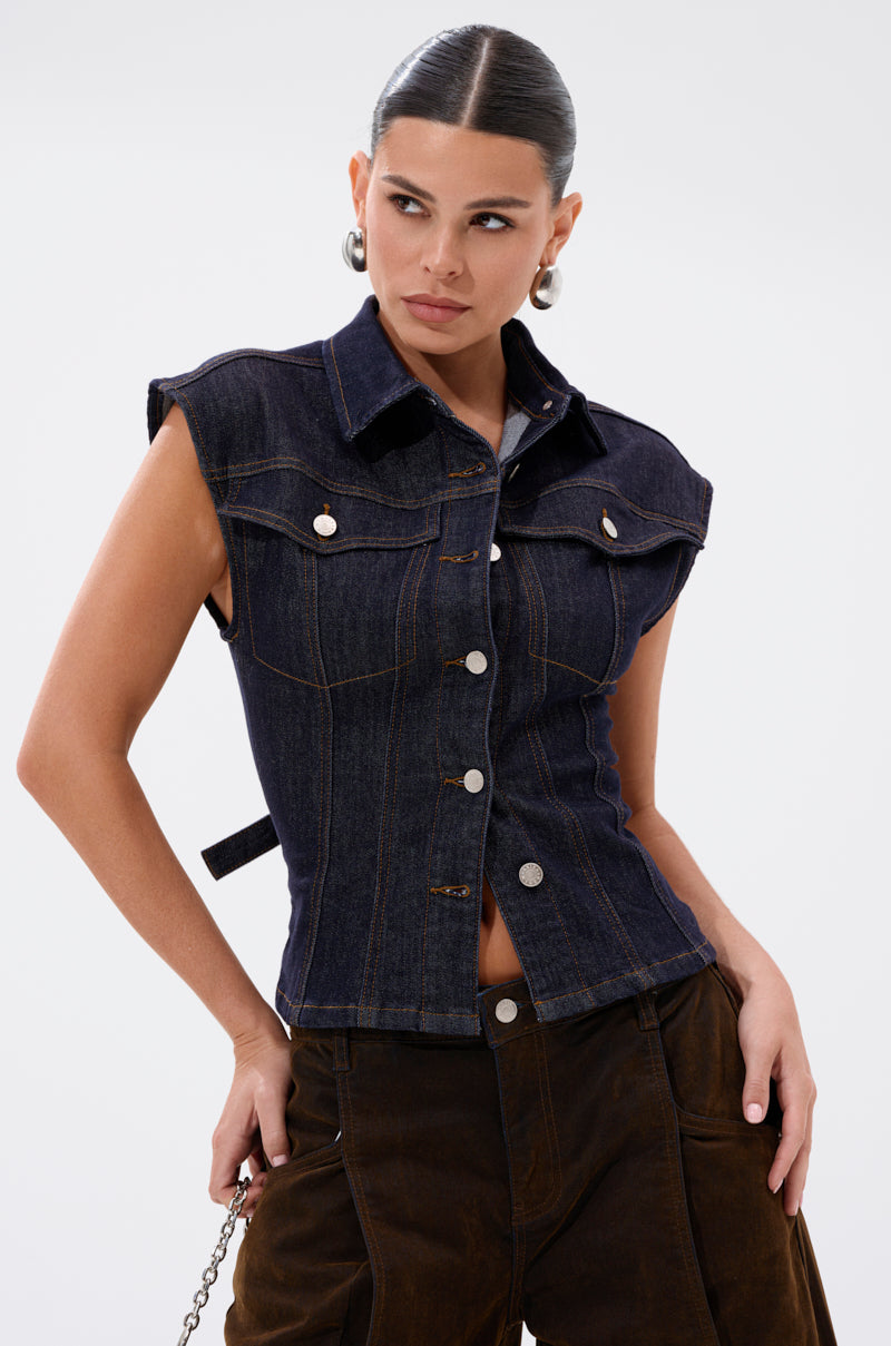 A woman poses with one hand on her hip against a white background, wearing the sleeveless LILY DENIM BUTTON DOWN vest featuring a dark-wash finish, large front pockets, and brown pants. 5