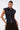 A woman poses with one hand on her hip against a white background, wearing the sleeveless LILY DENIM BUTTON DOWN vest featuring a dark-wash finish, large front pockets, and brown pants.