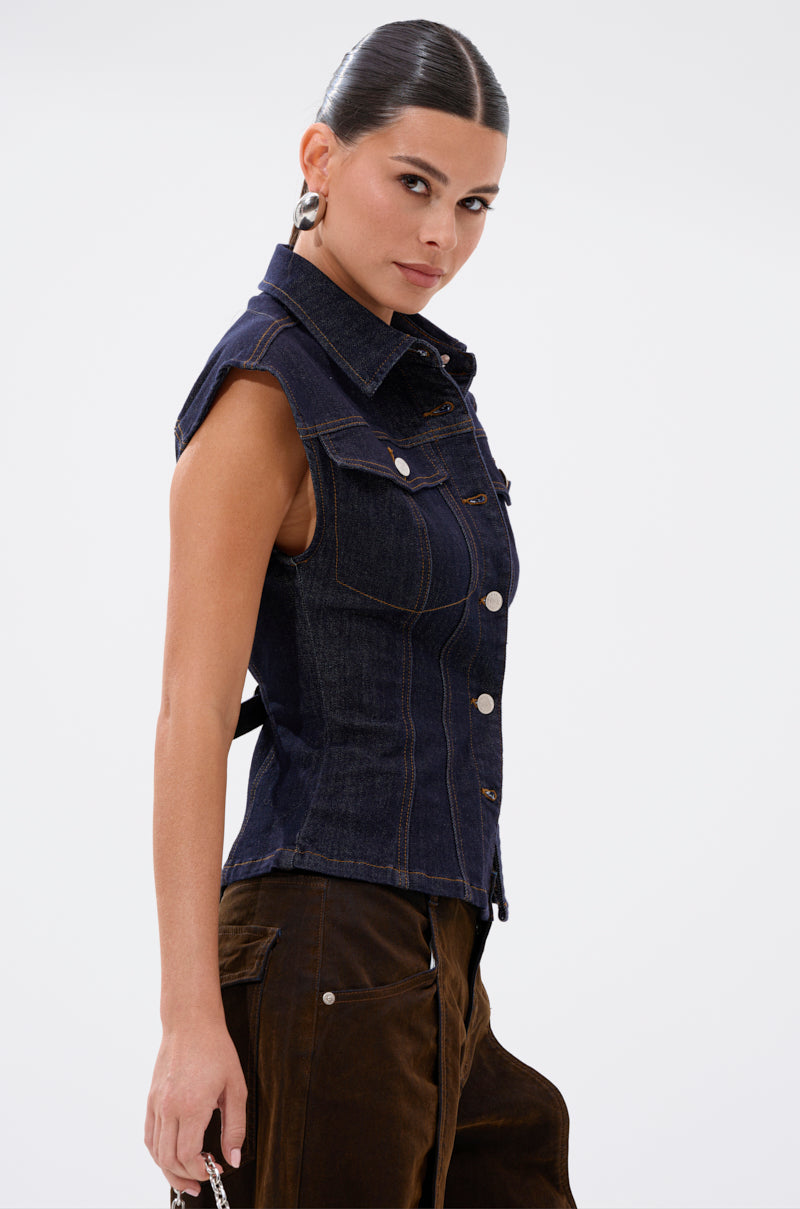 A woman with dark hair in a sleek bun models the LILY DENIM BUTTON DOWN—a form-fitting, sleeveless dark-wash denim vest—paired with brown pants. She stands in profile, looking over her shoulder against a plain white background. 6