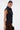 A woman with dark hair in a sleek bun models the LILY DENIM BUTTON DOWN—a form-fitting, sleeveless dark-wash denim vest—paired with brown pants. She stands in profile, looking over her shoulder against a plain white background.