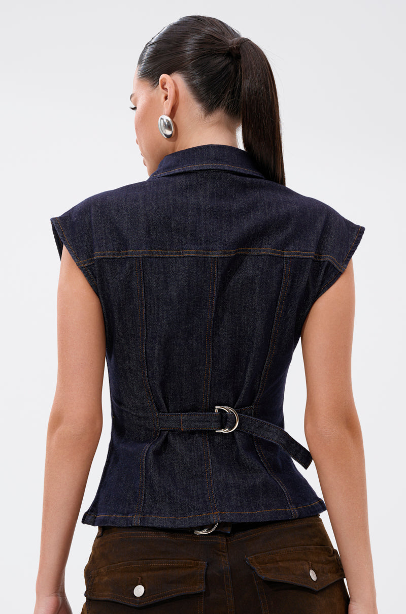 A woman with long dark hair in a ponytail wears the LILY DENIM BUTTON DOWN, a dark-wash cap-sleeve vest with a belted back, paired with brown pants with large back pockets. She faces away, highlighting the form-fitting silhouette. 7